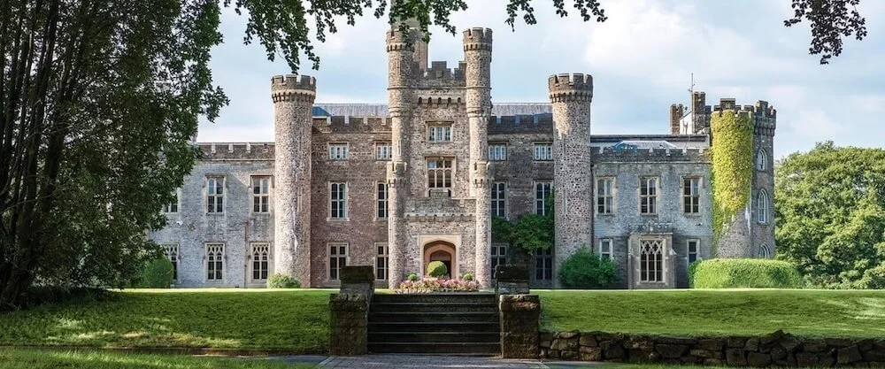 A large stone castle with multiple towers and turrets, surrounded by lush green trees and grass, with stairs leading up to the entrance.