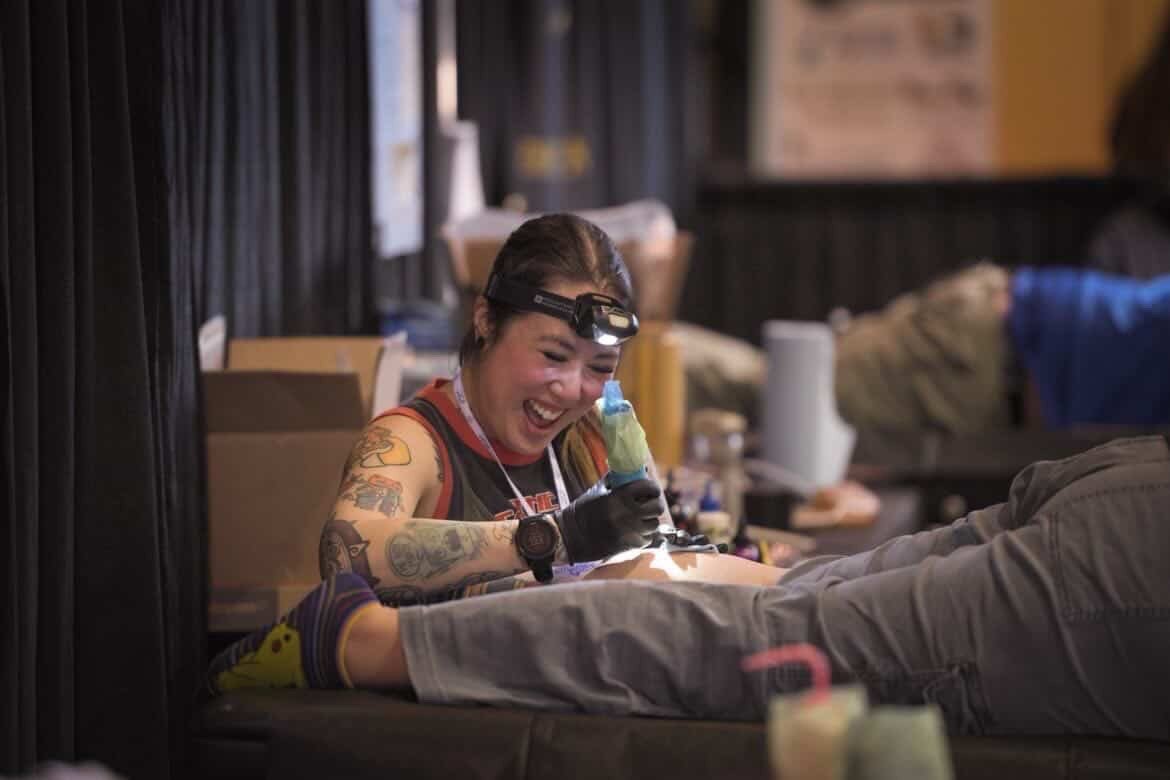 A woman tattoo artist, smiling with a headlamp, tattooing a person's leg in a tattoo parlor.