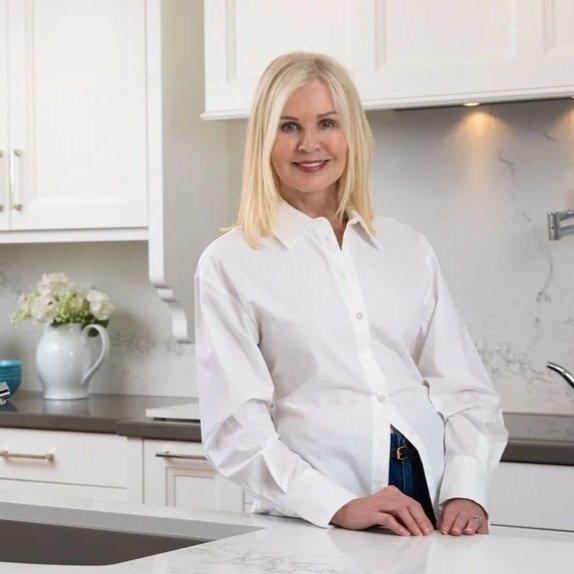 Jackie Glass in a white button-down shirt standing in a modern kitchen with white cabinets and marble countertops, smiling at the camera.