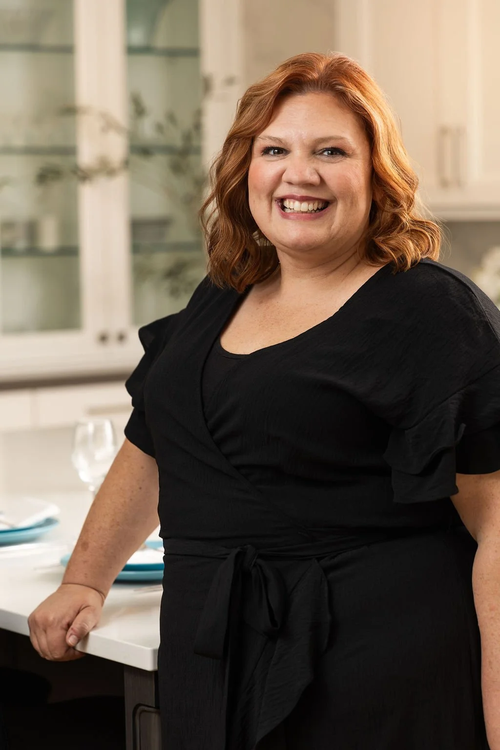 A smiling woman with red hair standing in a kitchen, wearing a black dress with ruffled sleeves.