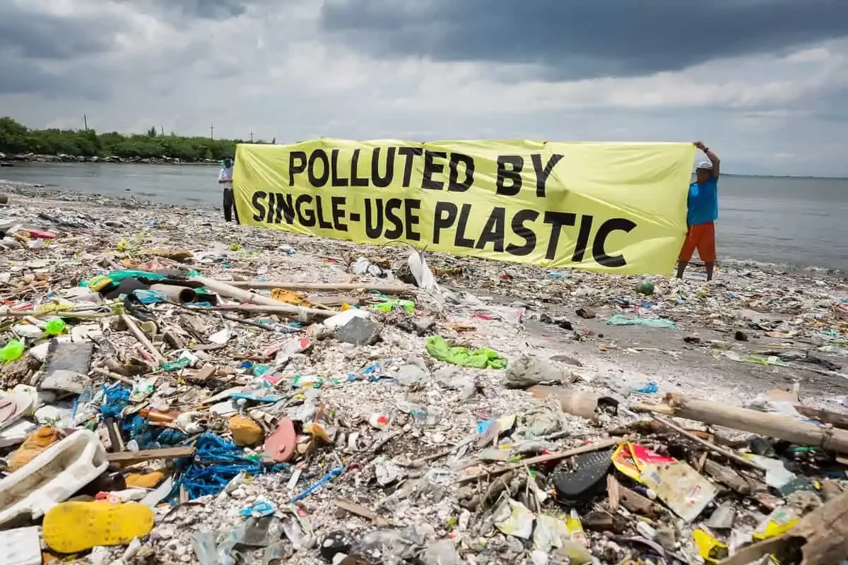 Beach filled with trash and debris, with two people holding a yellow banner that reads 'Polluted by single-use plastic', under cloudy sky.