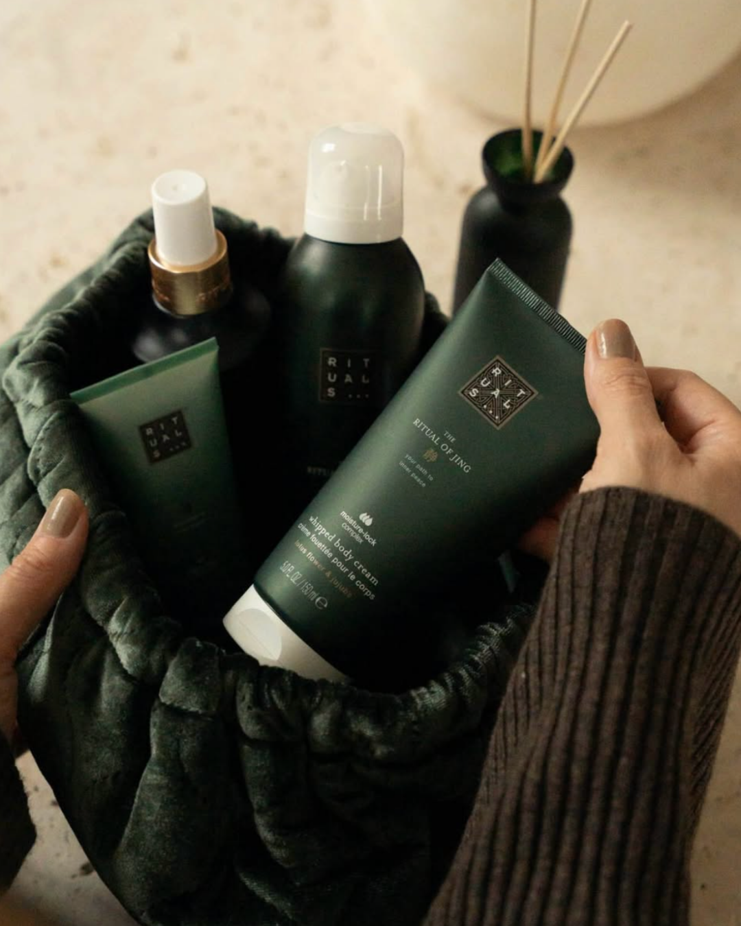 En person holder i hånden en grøn Rituals kropscreme, mens andre Rituals produkter ligger i en taske. Bagved er der en sort vase med små træpinde.