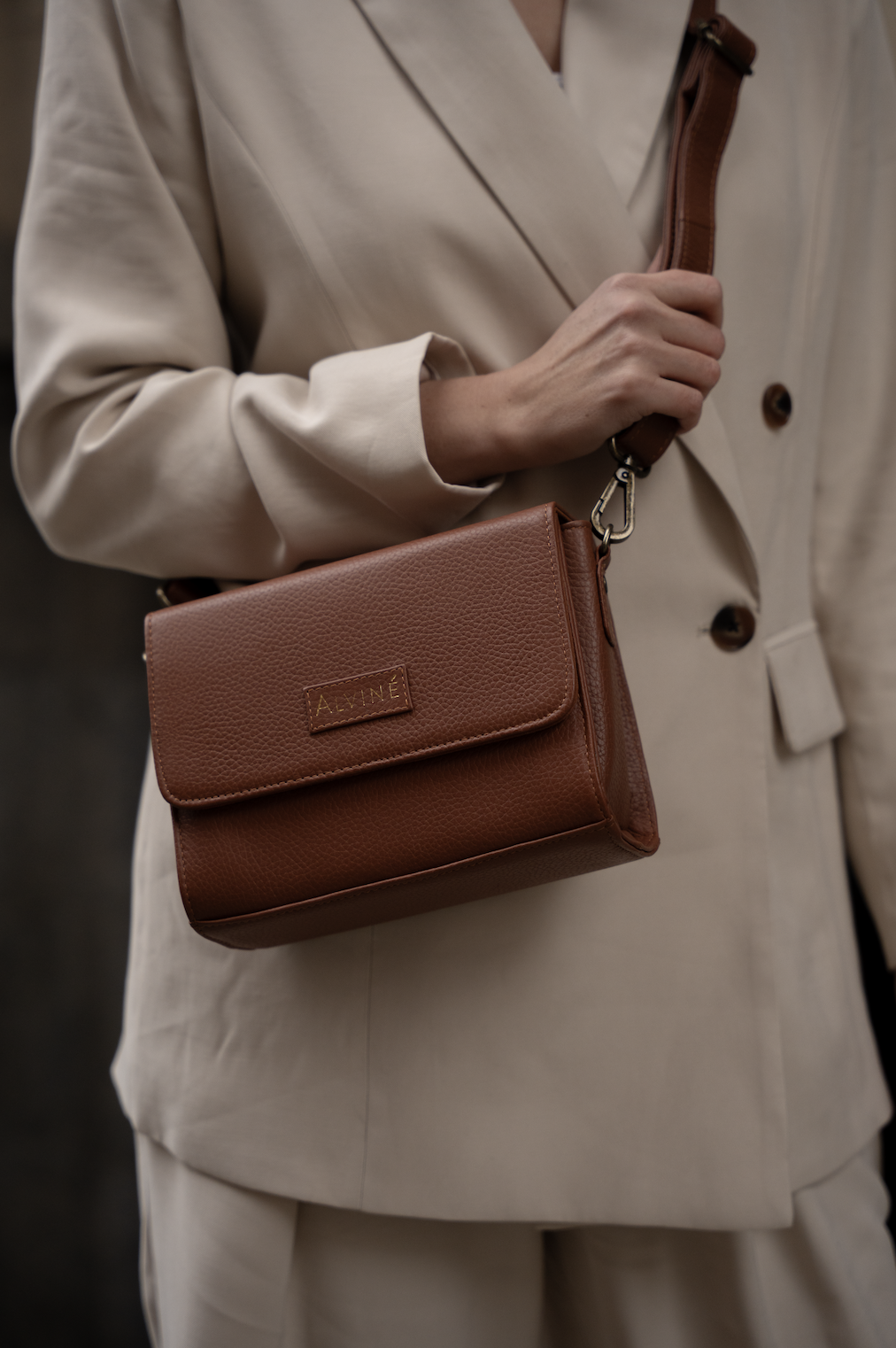 Person der bærer en beige frakke og en brun taske med navnet 'ALVINÉ' på, med fokus på taskens taske.