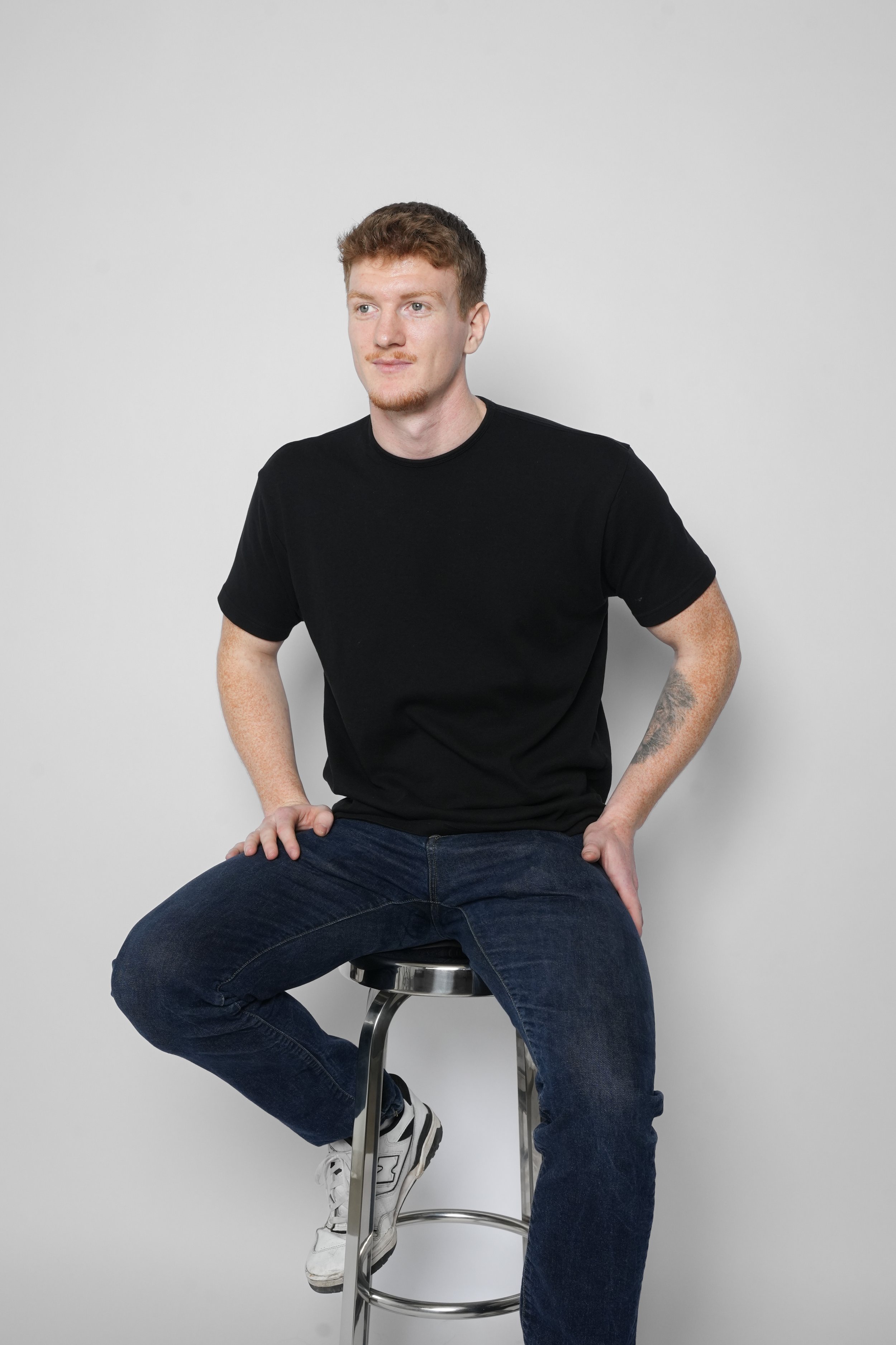Ung mand sidder på en barstol, iført sort T-shirt og blå jeans, foran en hvid baggrund.
