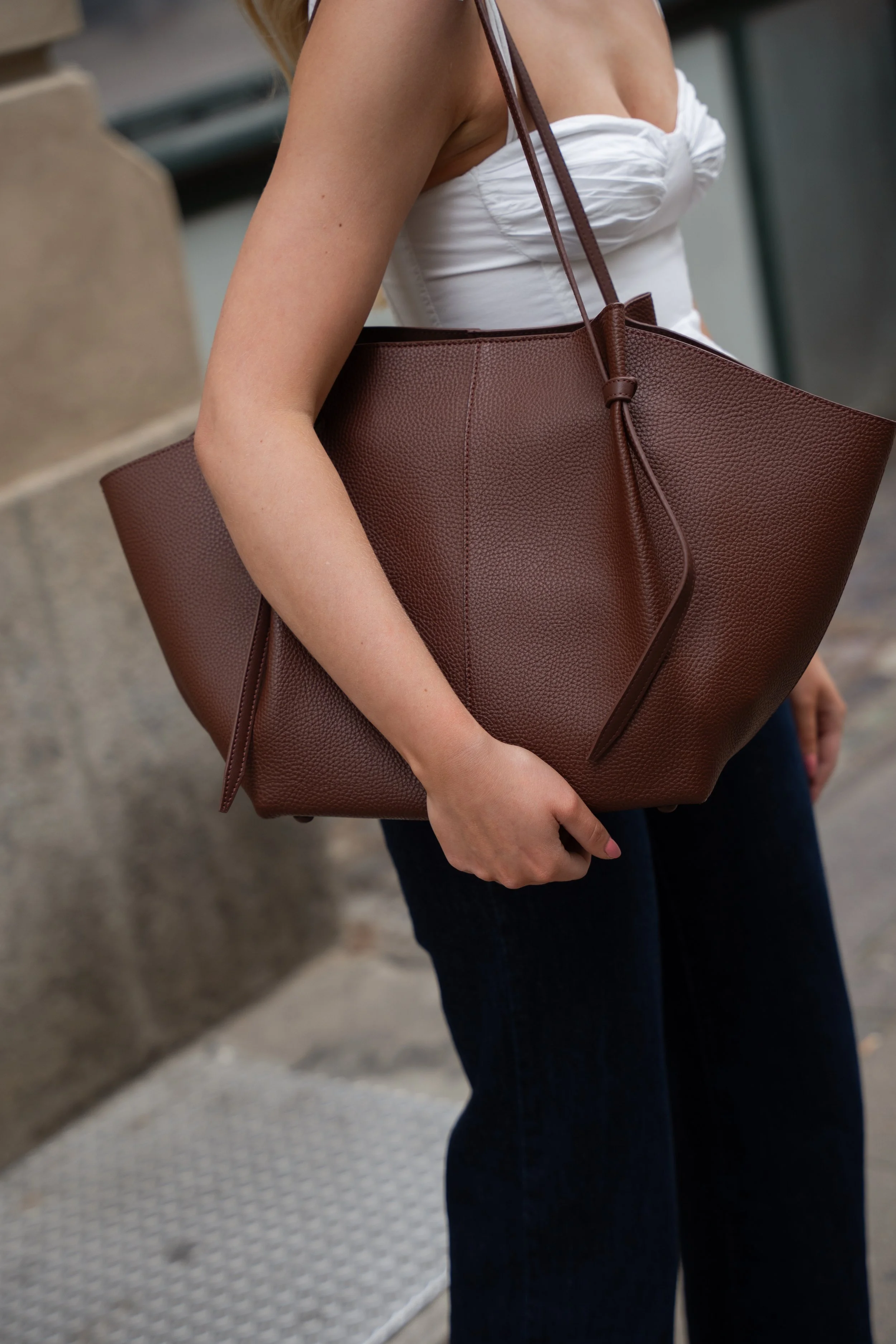 En person bærer en stor brun taske over skulderen, iført en hvid top og jeans, på en udendørs bygade.