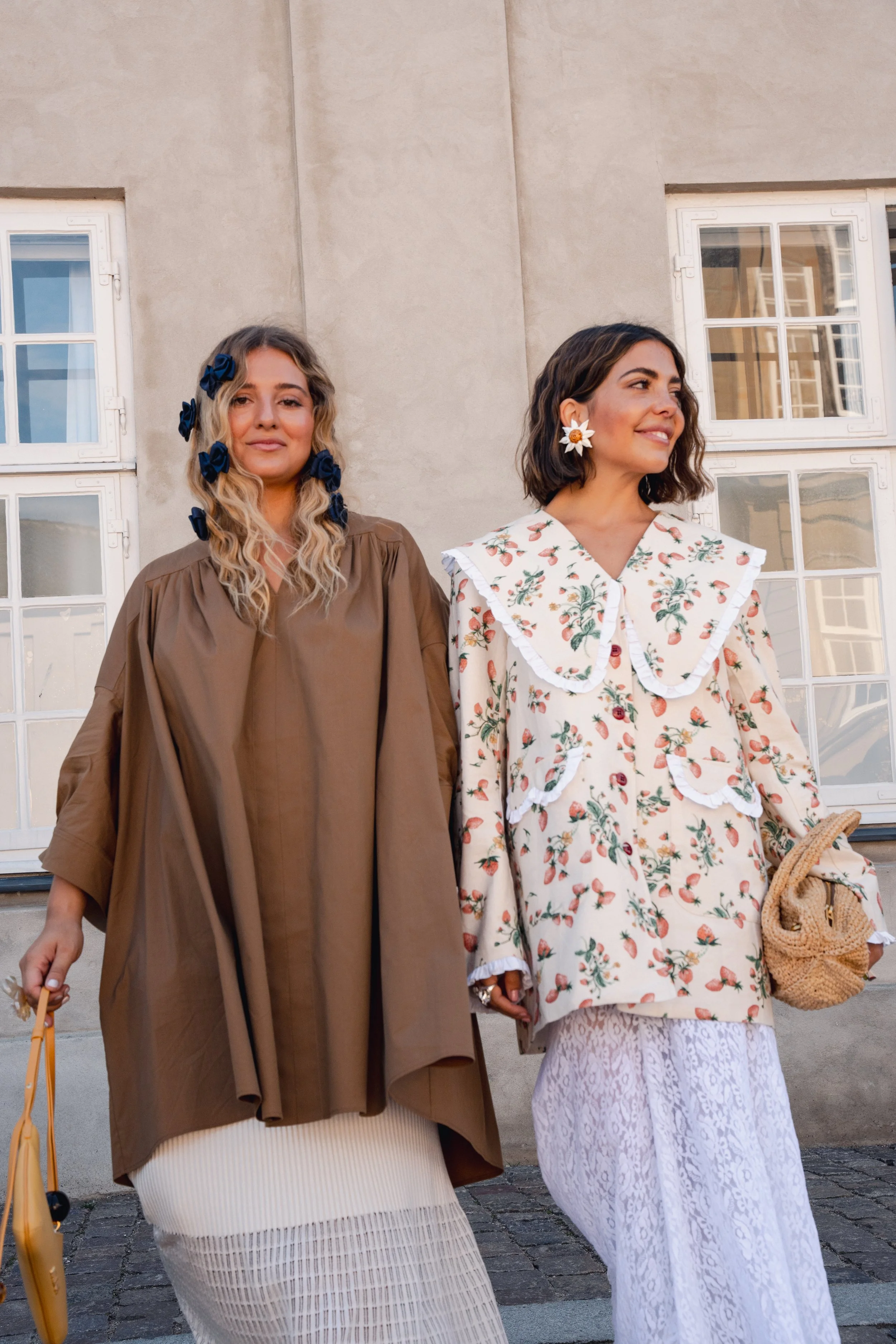 To kvinder går på gaden foran en bygning med hvide vinduer, iført flere lag tøj og accessoriseret med blomster i håret, en bærepose og en kurv.