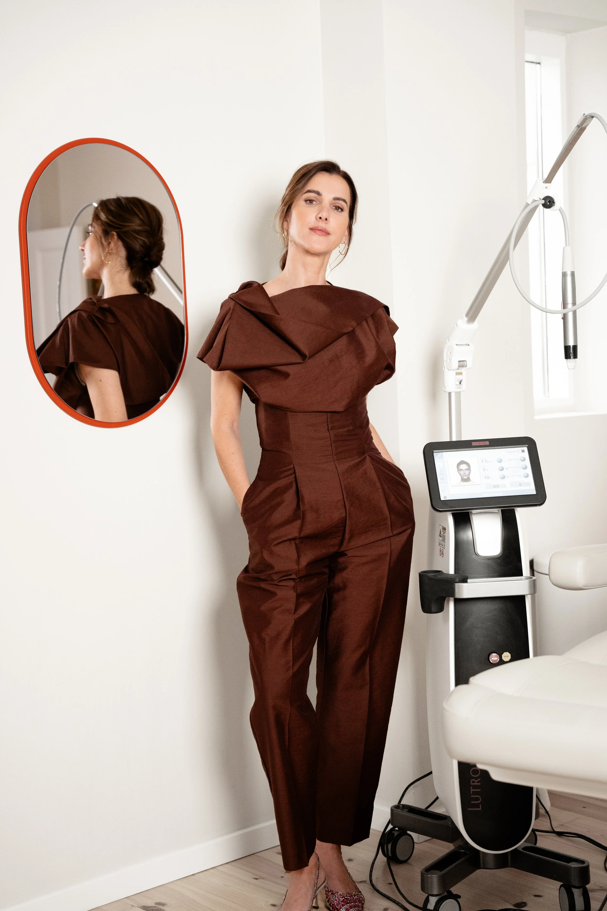 En kvinde står i et rum ved siden af en medicinsk maskine, med hænder i lommer, og kigger mod kameraet. Hun bærer en brun, designeragtig dragt med store skulderdetaljer, og der er et spejl bag hende, der reflekterer hendes profil.