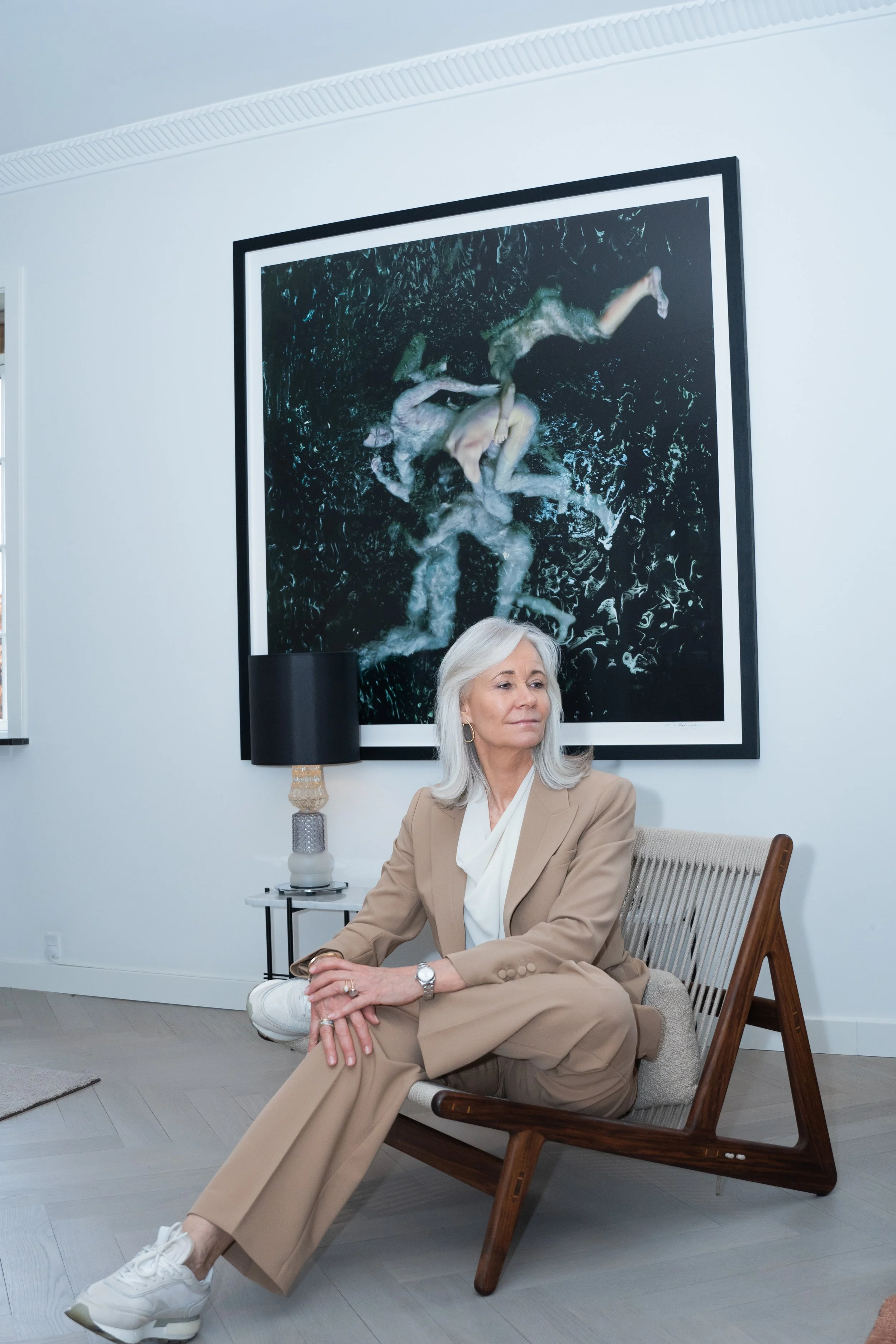 En ældre kvinde sidder på en stol i et lyst rum foran et stort kunstværk.