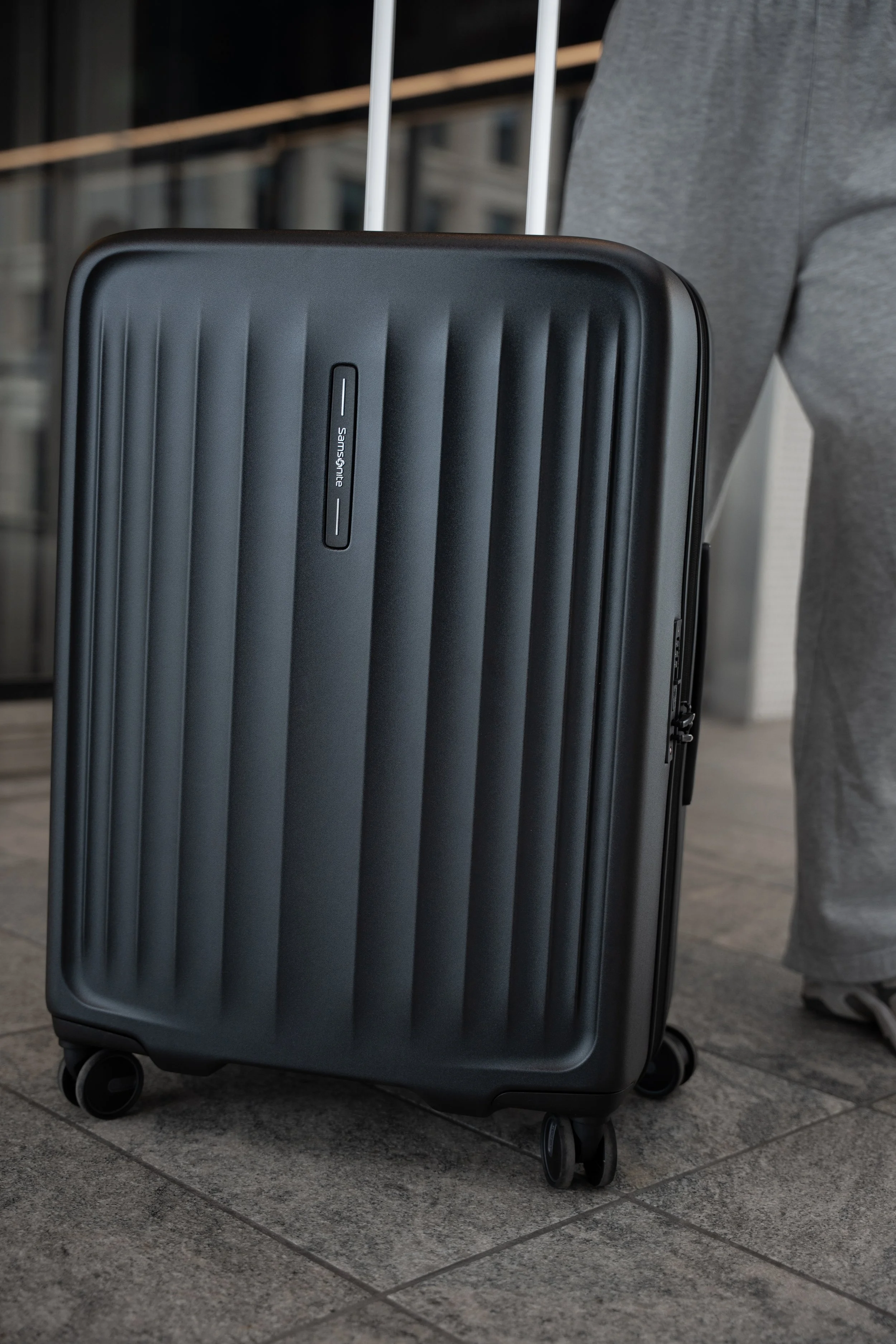 Sort hard shell kuffert med fire hjul, stående på en murstensbelagt fortov. 