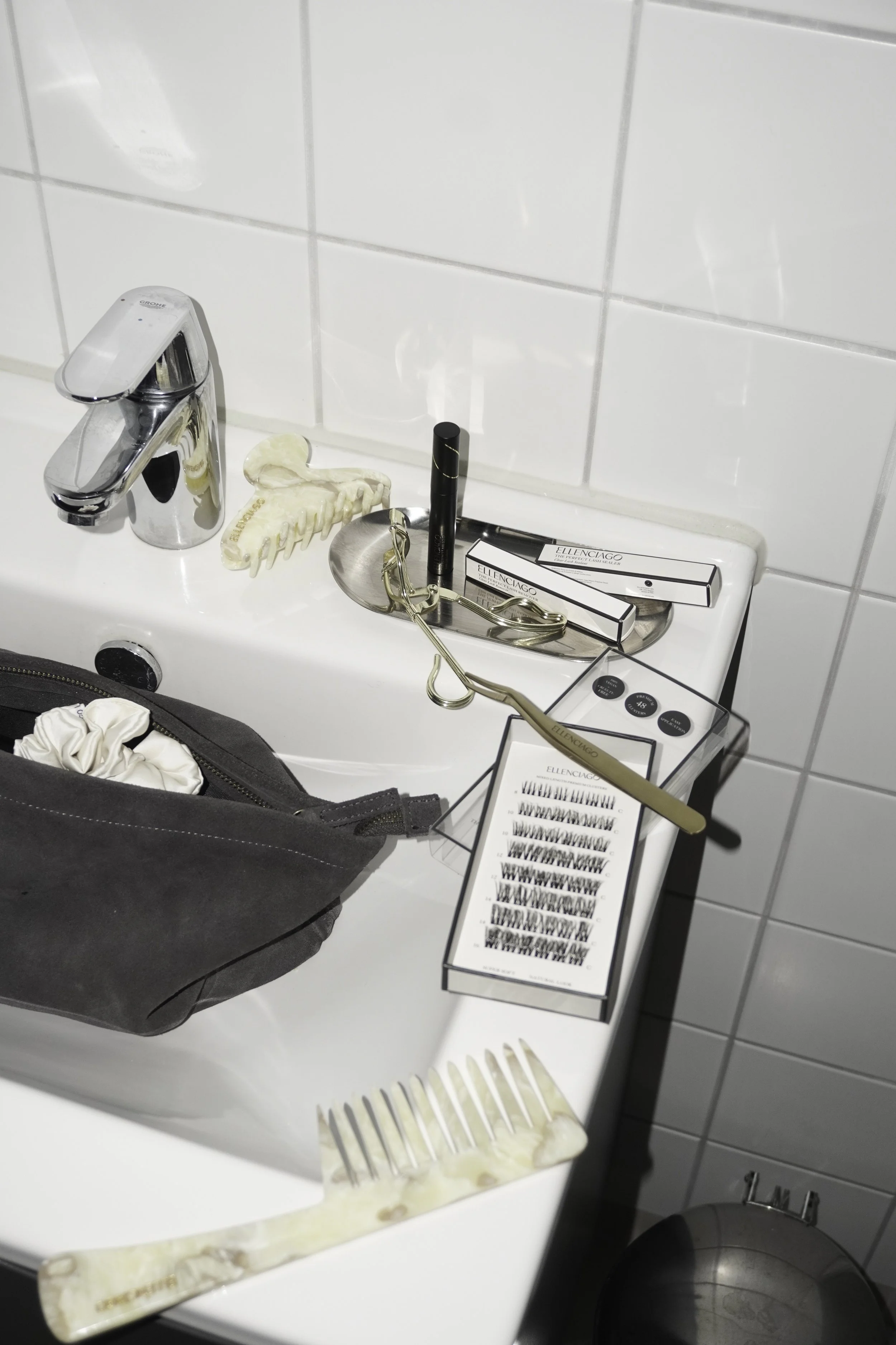 En vask med forskellige skønhedsprodukter, herunder øjenvipper, børste, øjenvippebukker og en makeuptaske, der er placeret på vasken.