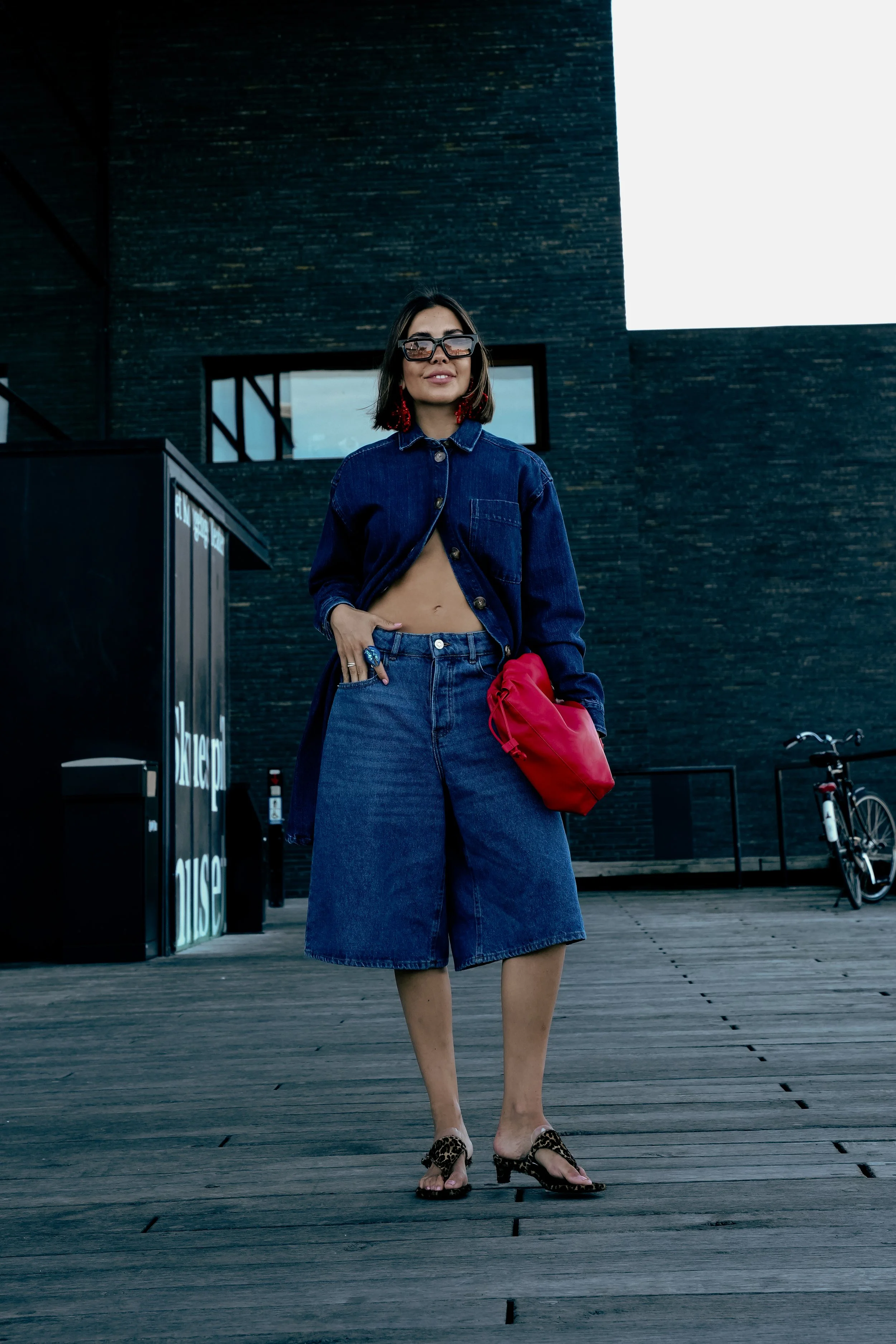 En kvinde står på en træbakke med en sort bygning i baggrunden, iført en denimjakke, nederdel, høje hæle og store solbriller, og holder en rød taske.