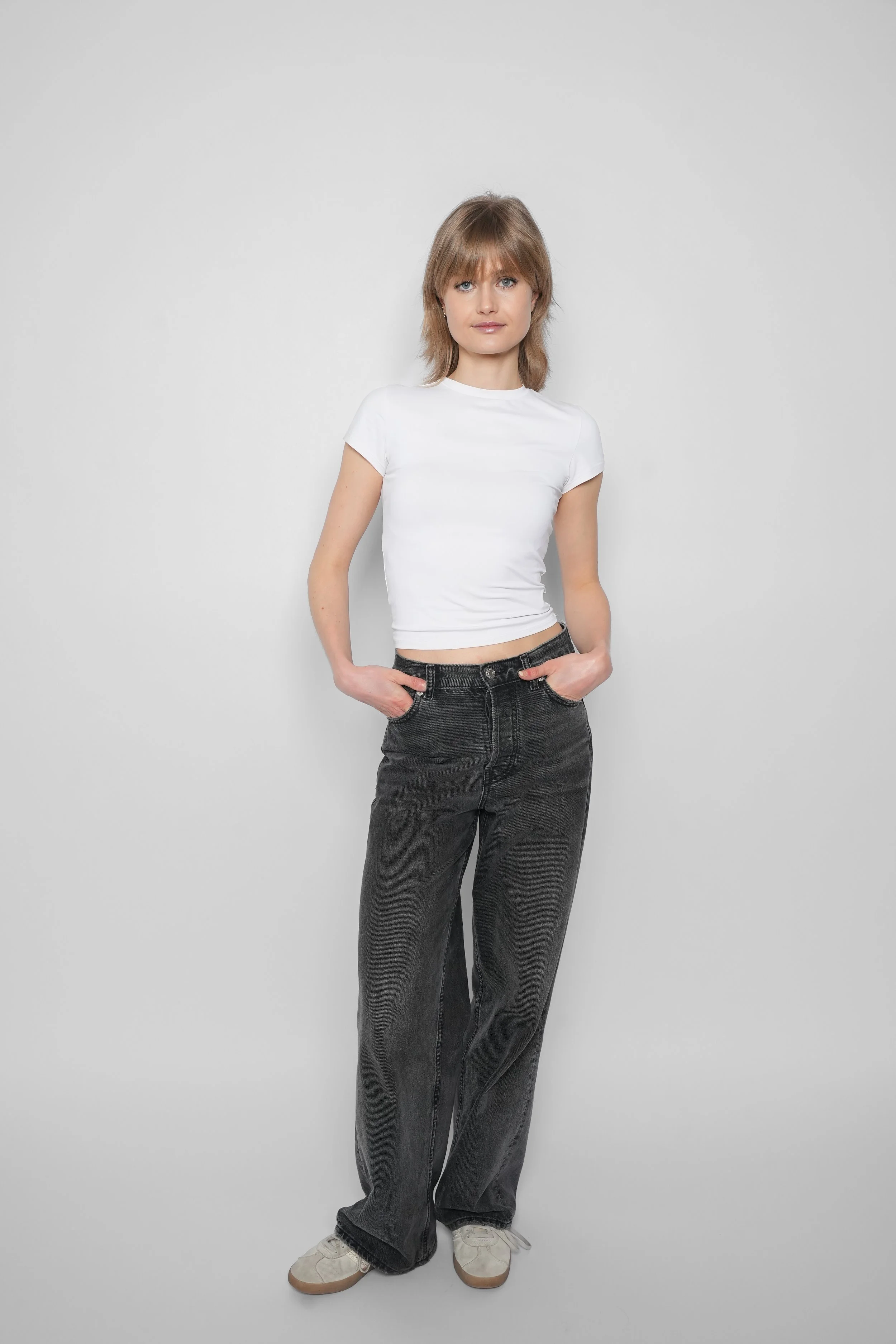 En ung kvinde står ved en blank, hvid baggrund, iført en hvid T-shirt og løse, mørke jeans.