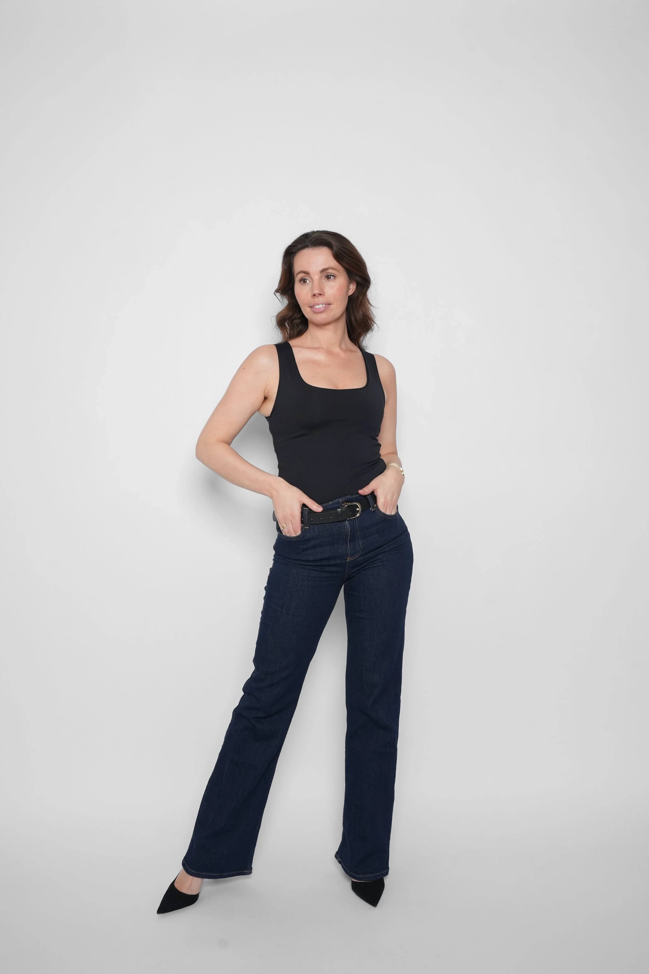 Kvinde iført sort top, blå jeans og sorte sko, stående mod en hvid væg.