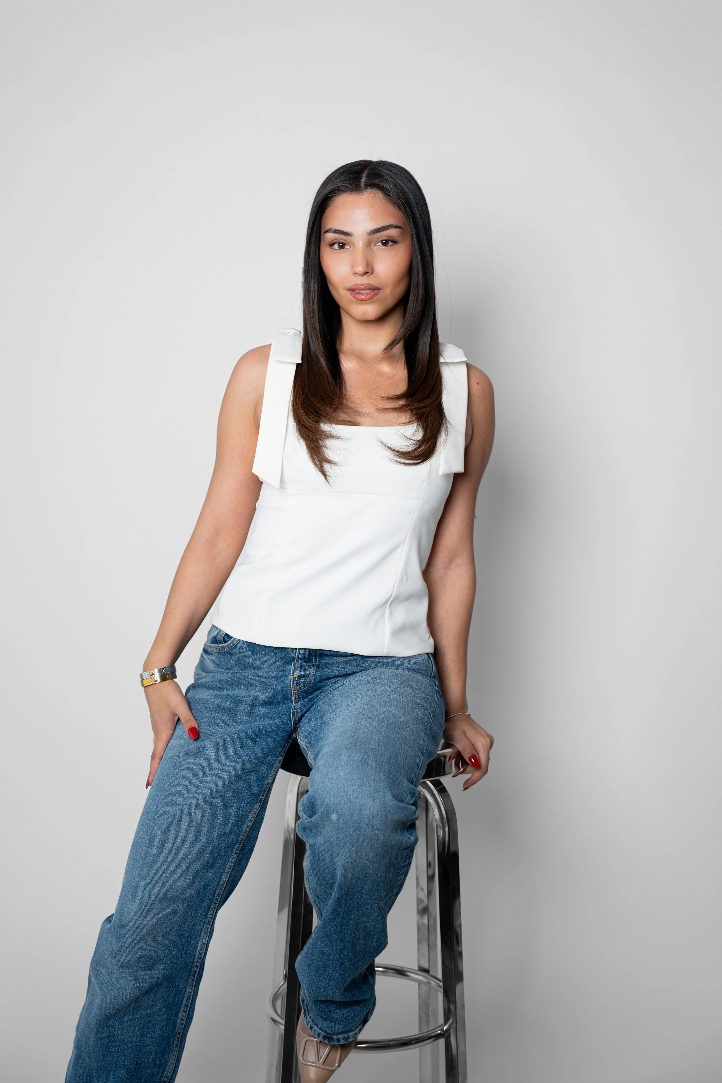 En ung kvinde med mørkt, lige hår, iført hvid top og blå jeans, sidder på en barstol mod en hvid væg.