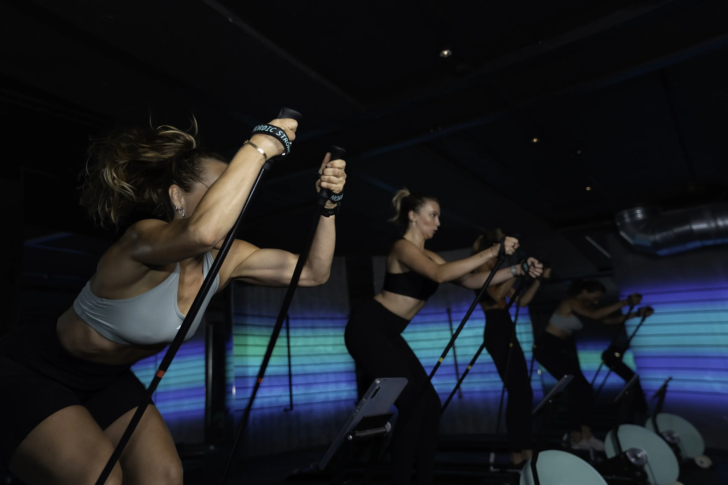 Kvinder der træner på en maskine med stænger, træner i et mørkt fitnesscenter med blå og grønne lysfacetterede vægge.