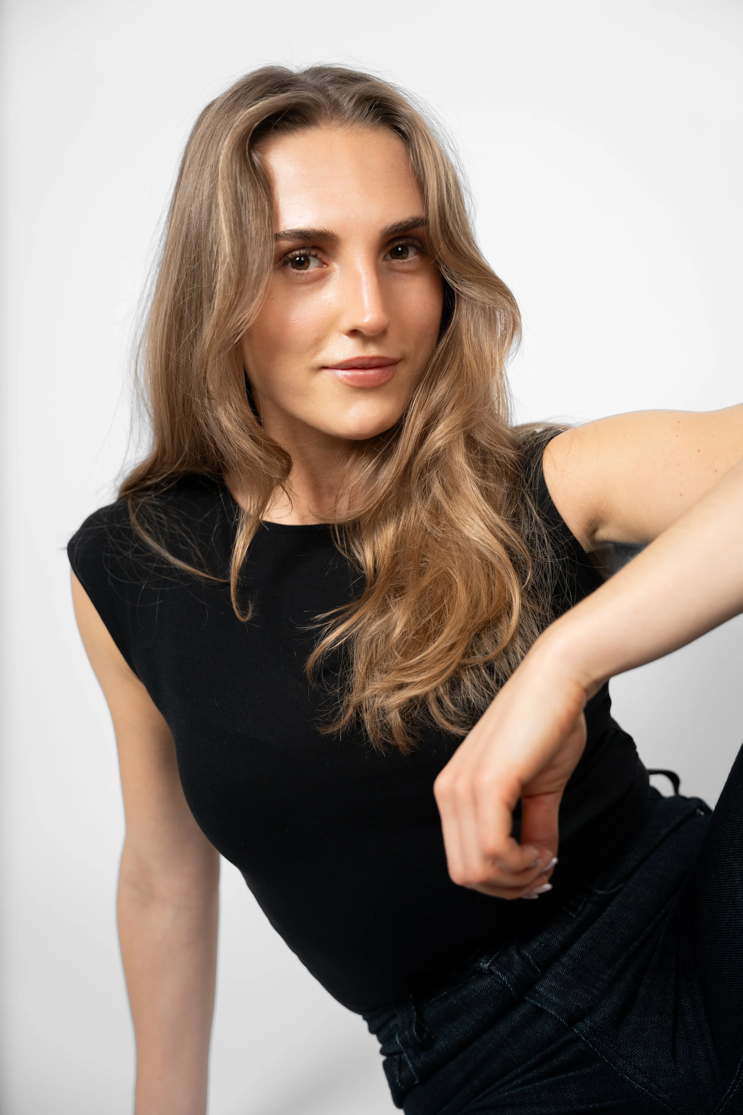 En ung kvinde med lang, bølget, lys brun hår, iført sort t-shirt, poserer mod en hvid baggrund.