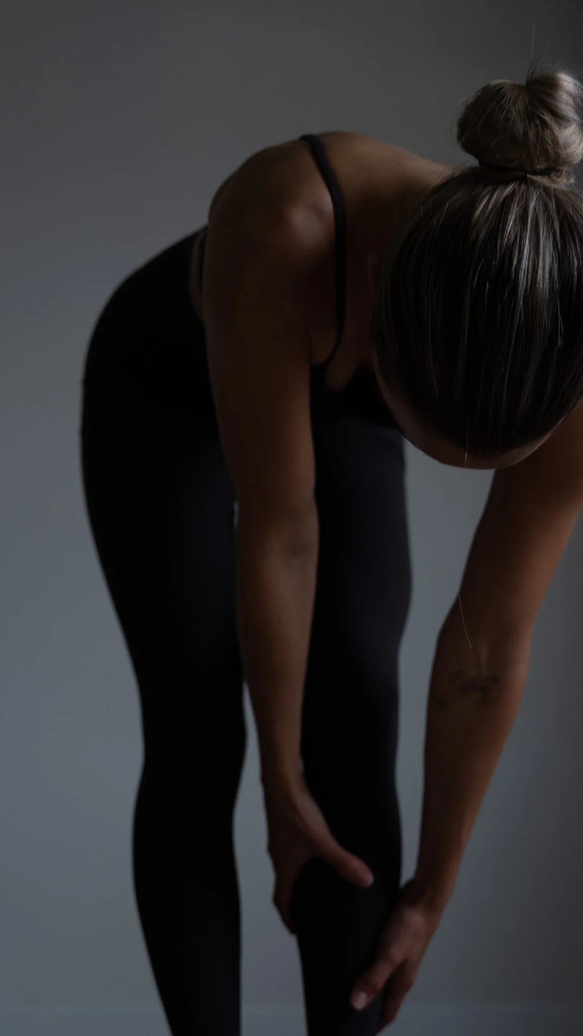 En kvinde bøjer sig forover, holder i sit højre ben, i sort træningstøj, med lyst hår i en knold.