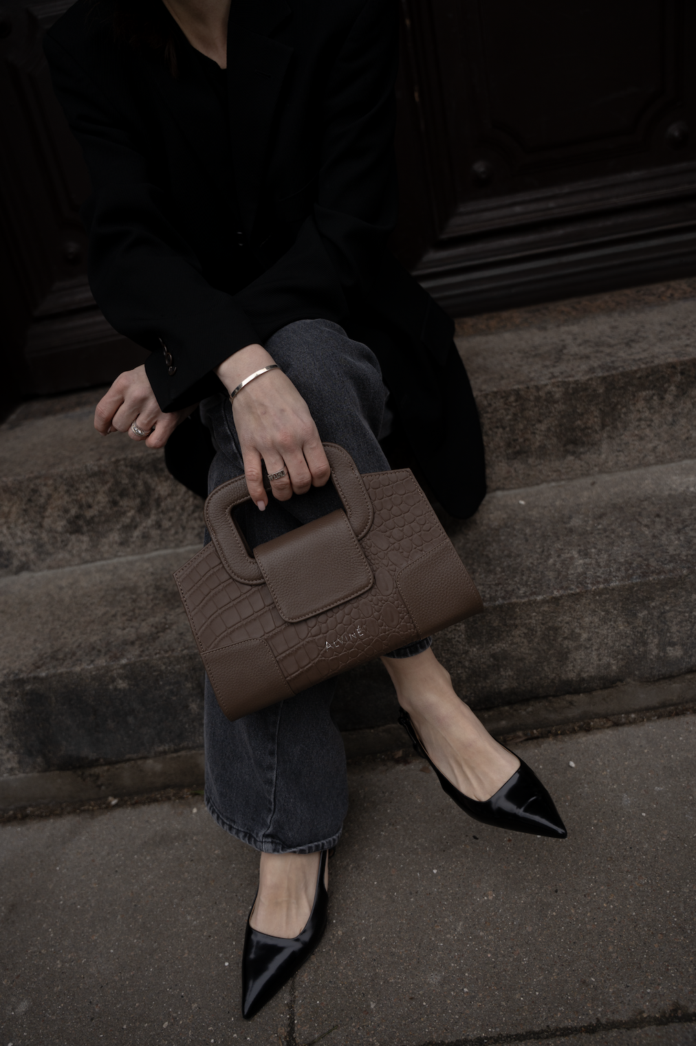 En person sidder på en trappe iført sort jakke, grå bukser og sorte stiletter, og holder en brown taske.