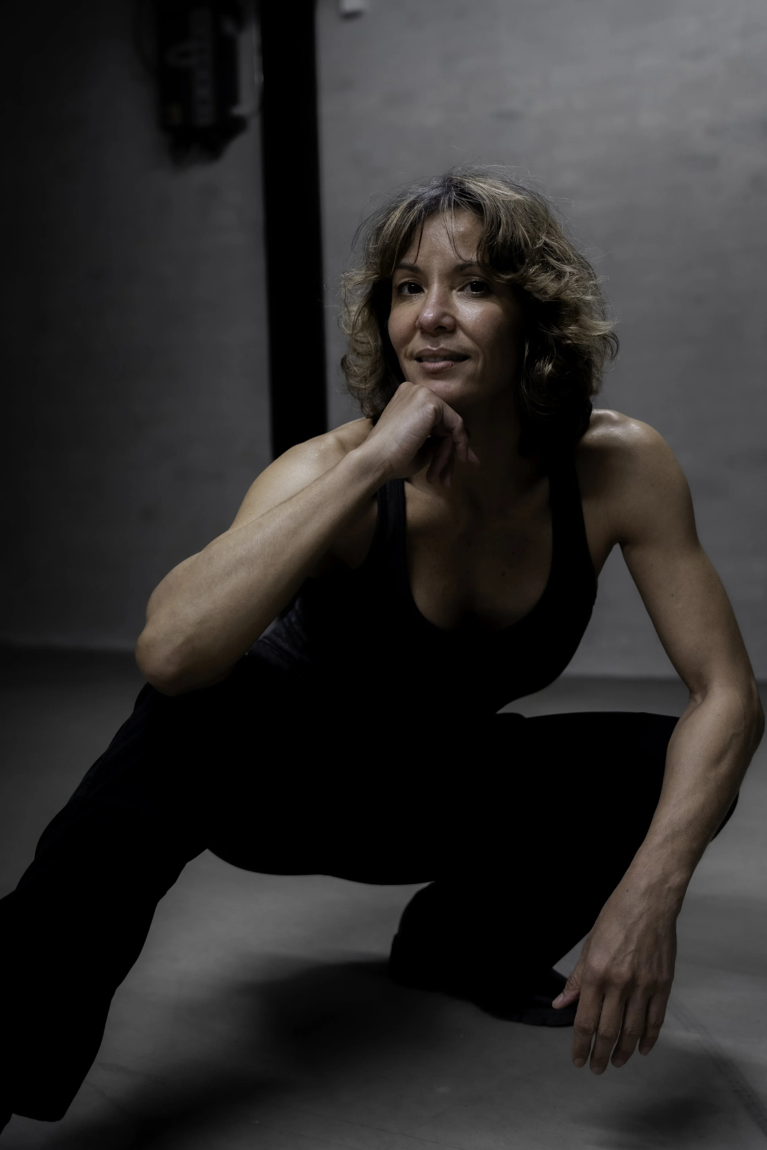 Kvindelig person med brunt, krøllet hår i sort top, squats ned på gulvet i et industrielt rum med grå vægge.