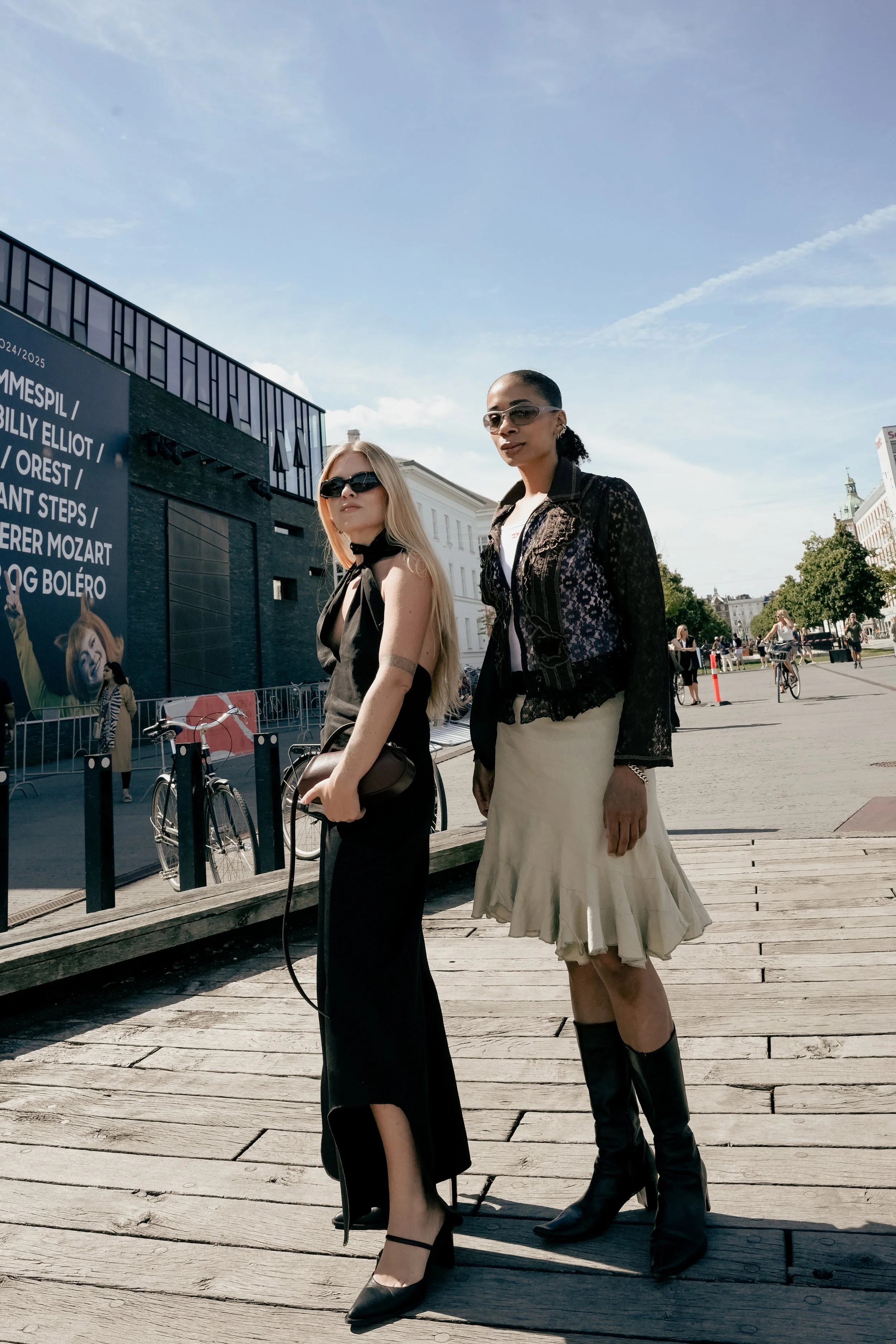 To kvinder står på en træpromenade udendørs, iført moderigtige tøj med solbriller. En er blond og bærer en sort kjole, mens den anden har mørk hud, sort hår, en sort blondejakke og en beige nederdel.