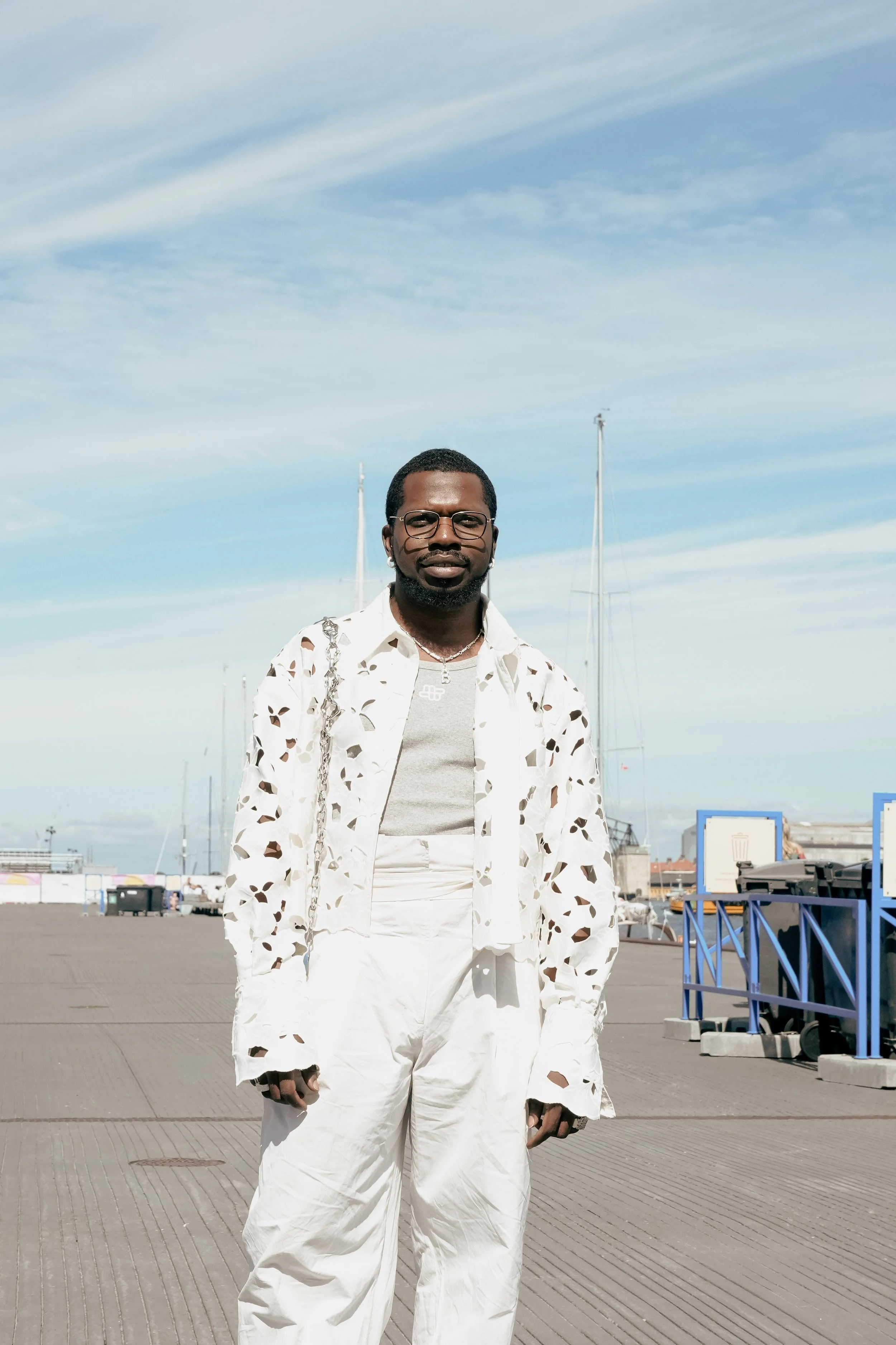 Mand står på en marina med både og bådlys i baggrunden under en blå himmel med spredte skyer.