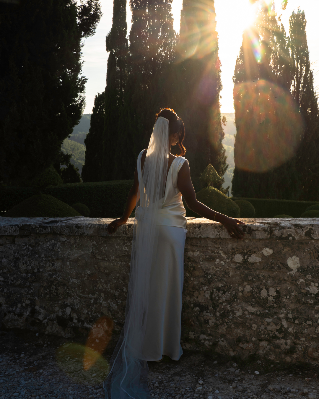 En kvinde i en hvid kjole med slør, der står med ryggen mod kamera foran en stenmur, omgivet af store træer, med sollys, der skinner gennem træerne.