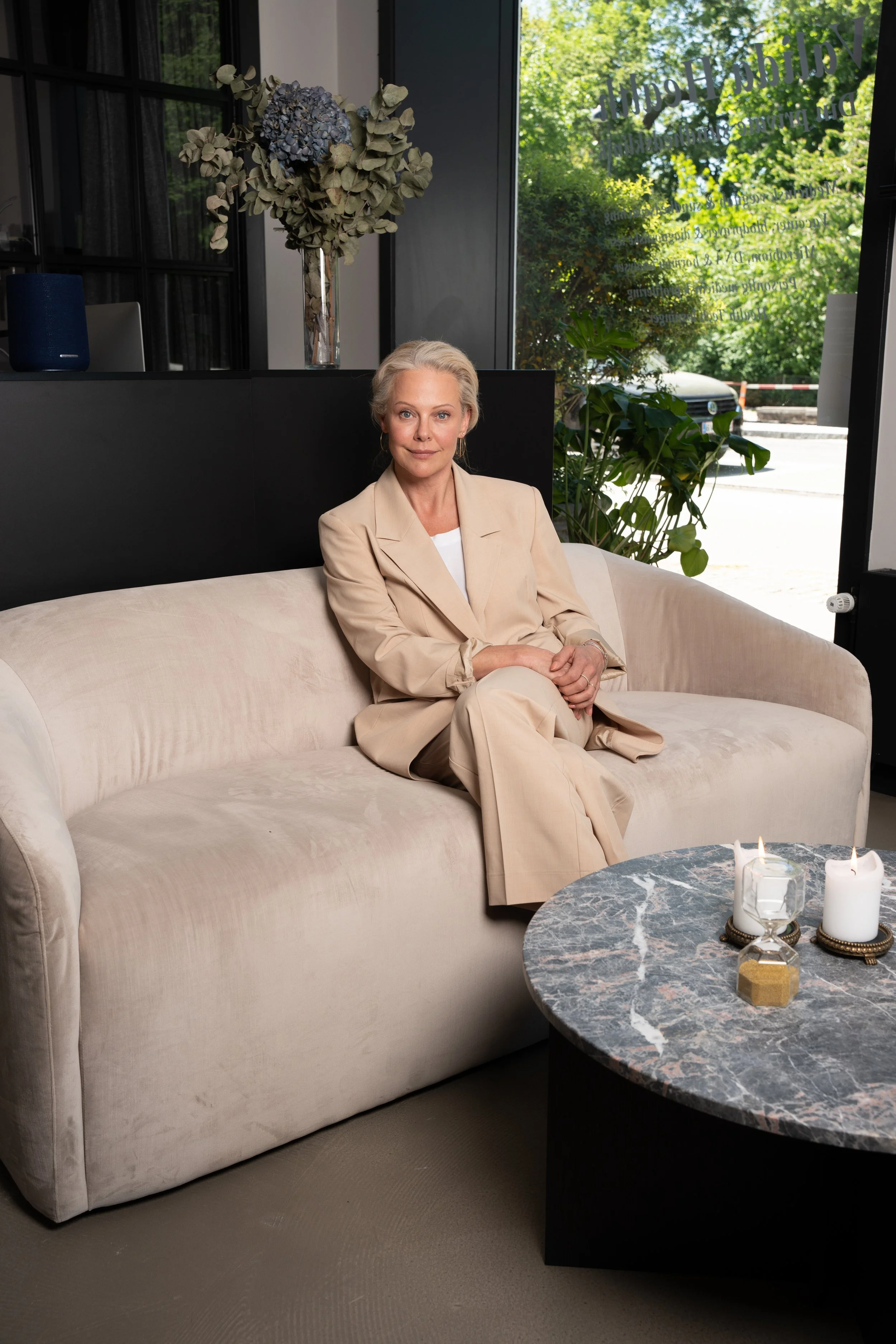 En ældre kvinde sidder på en beige sofa i et moderne rum med store vinduer. Der er en dekoration med tørret blomster foran vinduerne og stearinlys på et sort marmorbord.