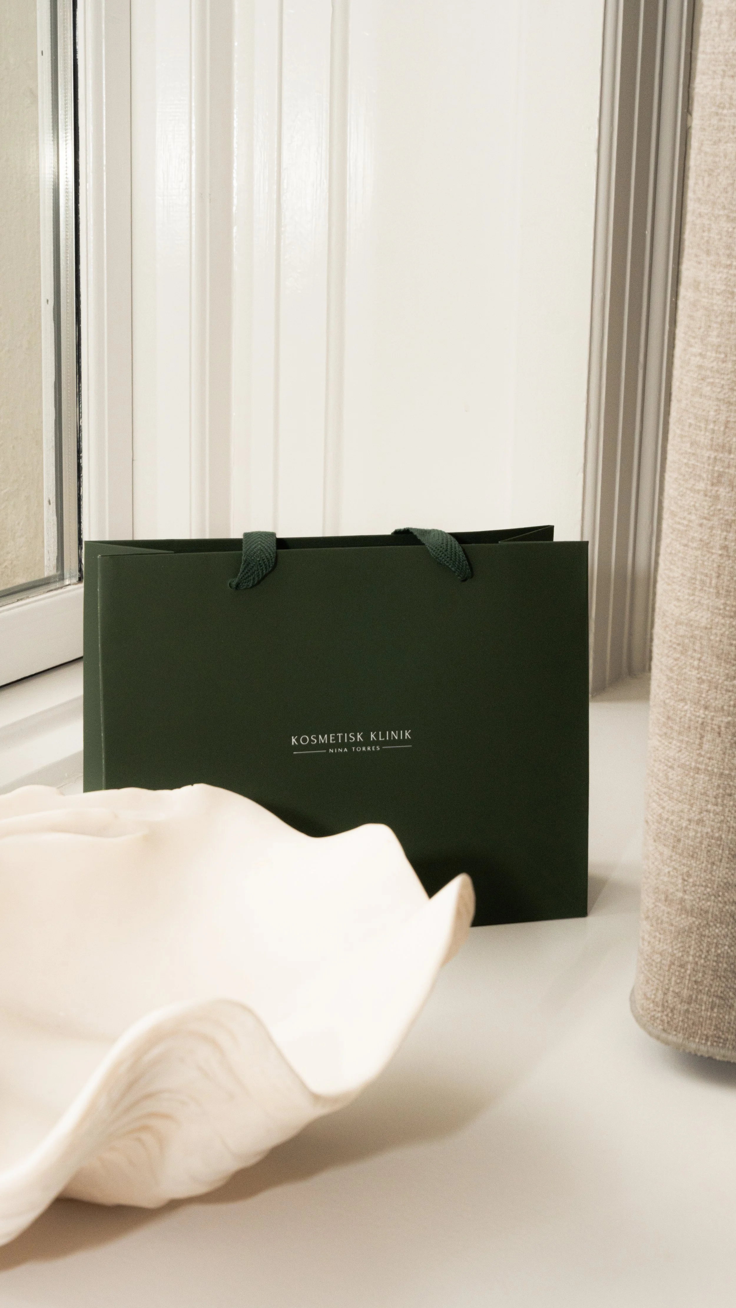 En grøn pose med teksten "KOSMETISK KLINIK NINA TORRES" står på en hvid overflade, med en stor muslingeskal foran.