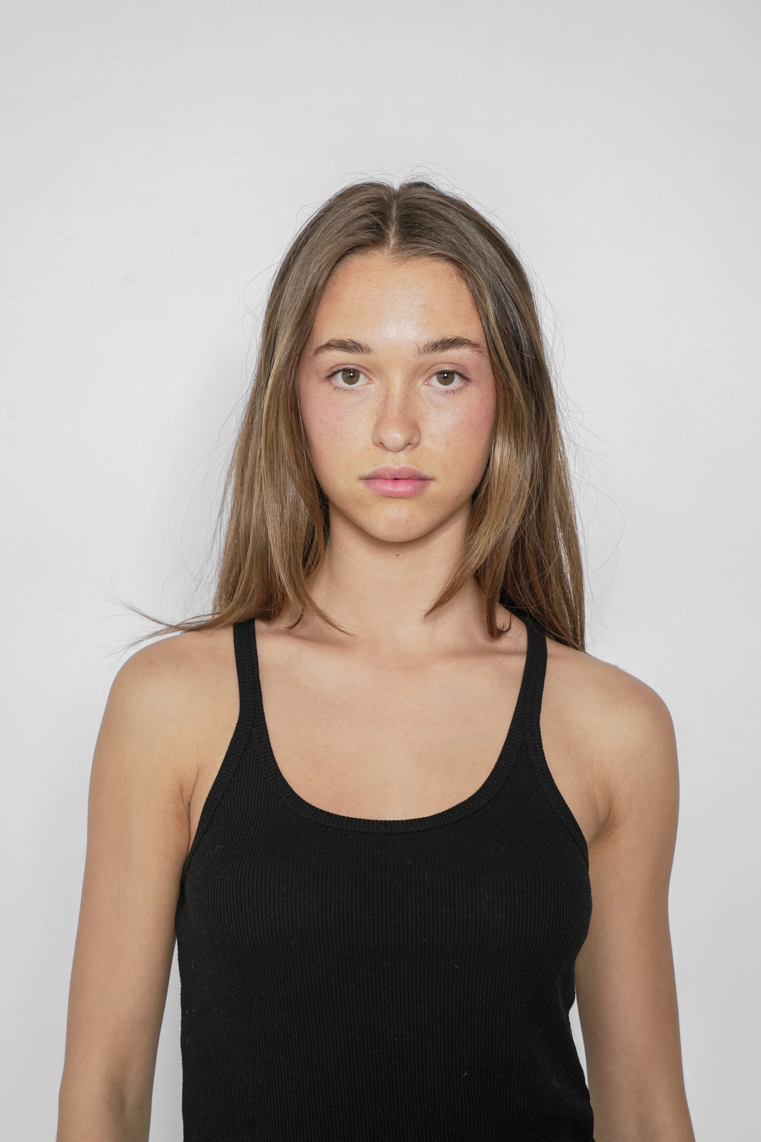 Portræt af en ung kvinde med brunt, langt hår, iført en sort top, stående foran en hvid baggrund.
