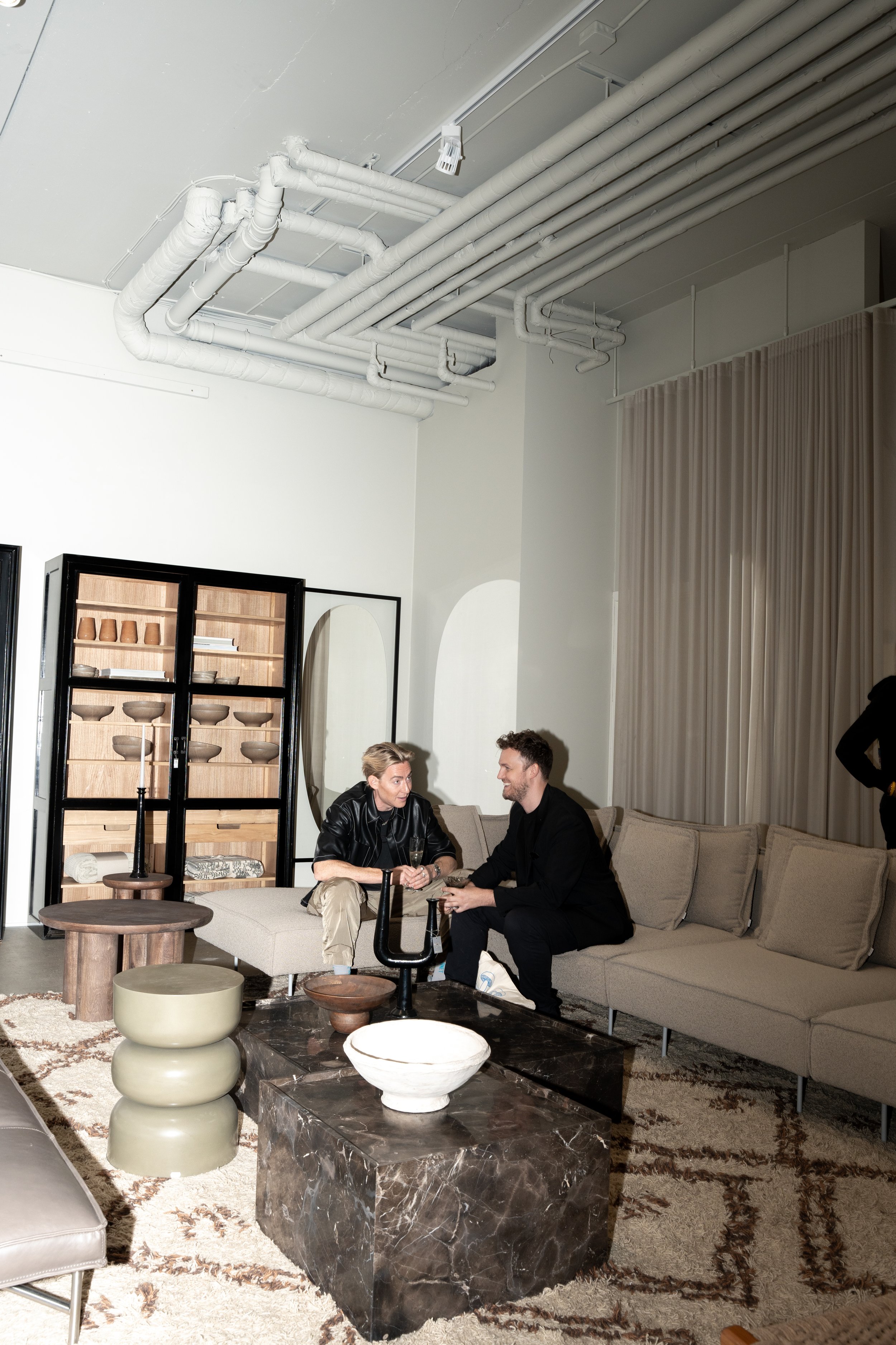 To mennesker sidder på en sofa og taler i et lækkert stueområde med moderne møbler og dekor. Der er synlige loftrør og en stor spejl i baggrunden.