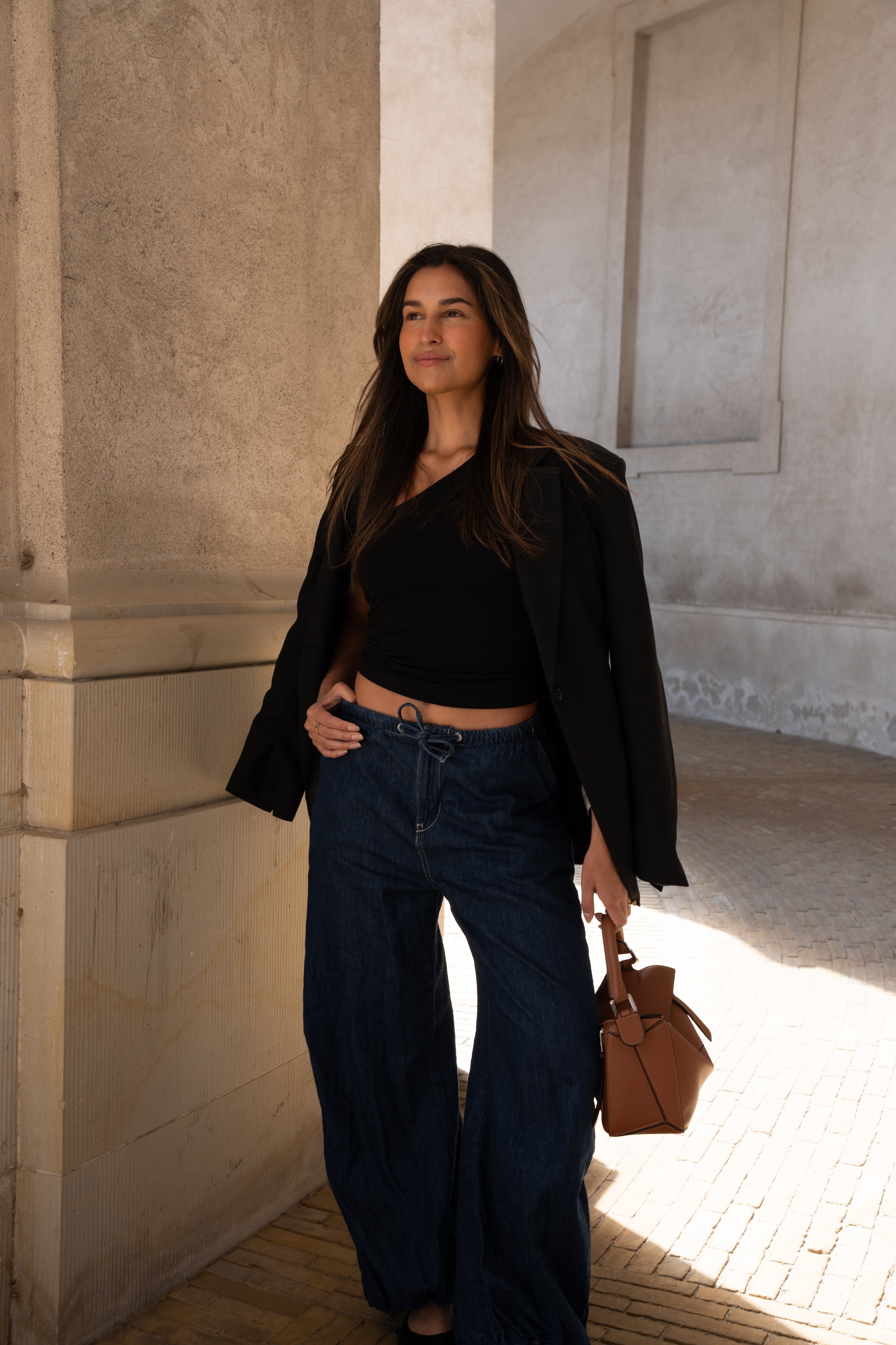 En kvinde iført sort top, blazer og løse jeans står i en arkitektonisk bygning med betonvægge og stenbelægning.