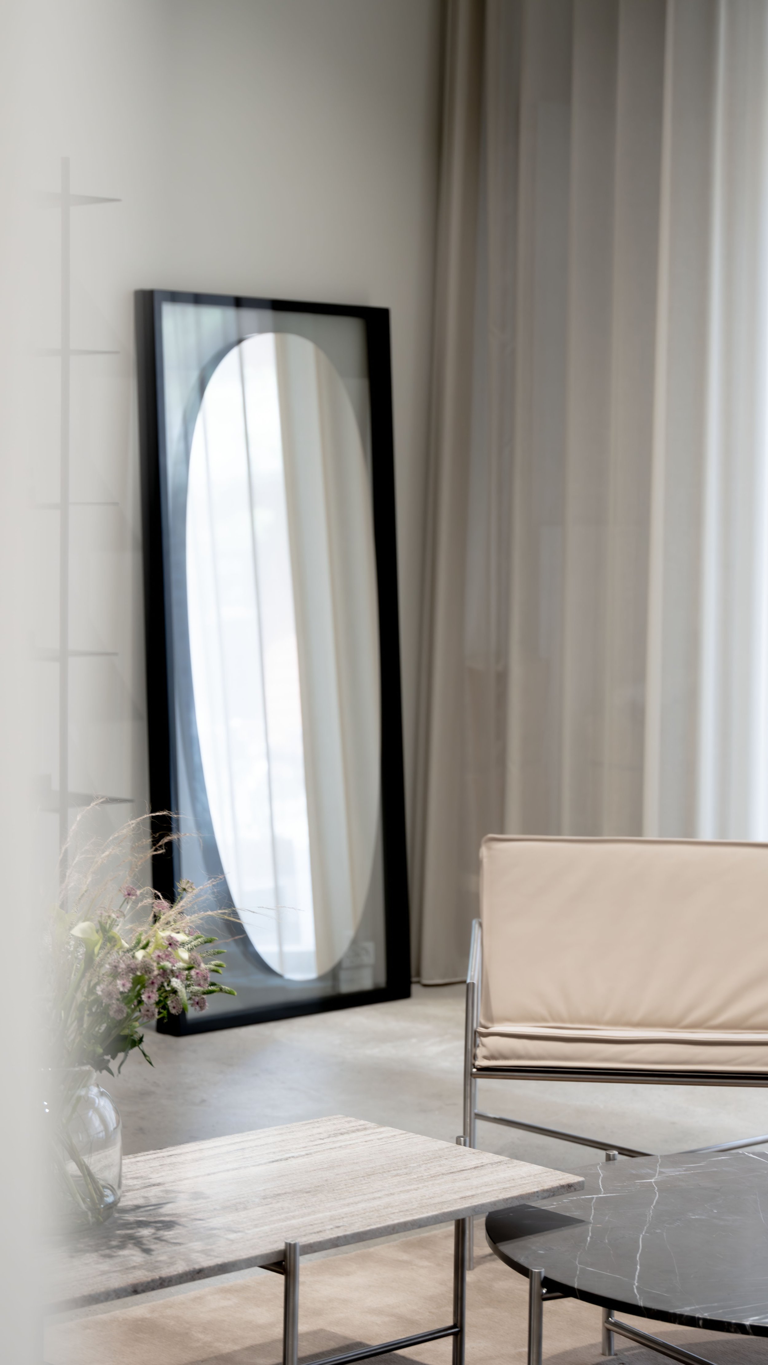 En stor, sort ramme spejl står mod en væg i et lyst, moderne rum med beige gardiner, en beige lænestol, et marbleborde og en vase med blomster.