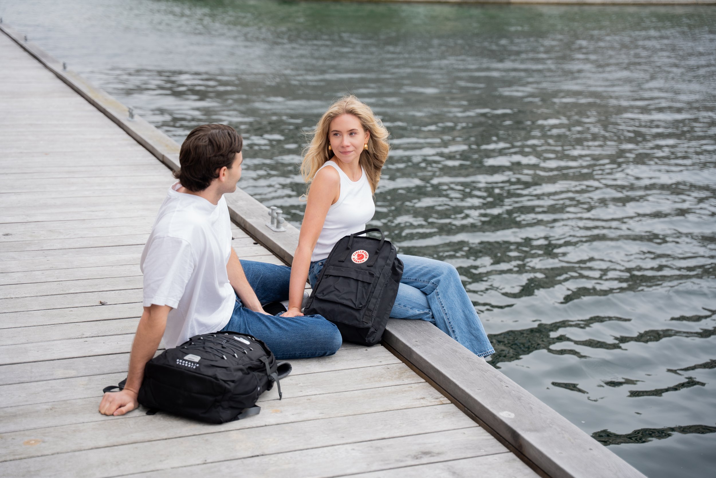 To unge voksne sidder på en træpier ved vandet med rygsække foran sig, og de taler med hinanden.