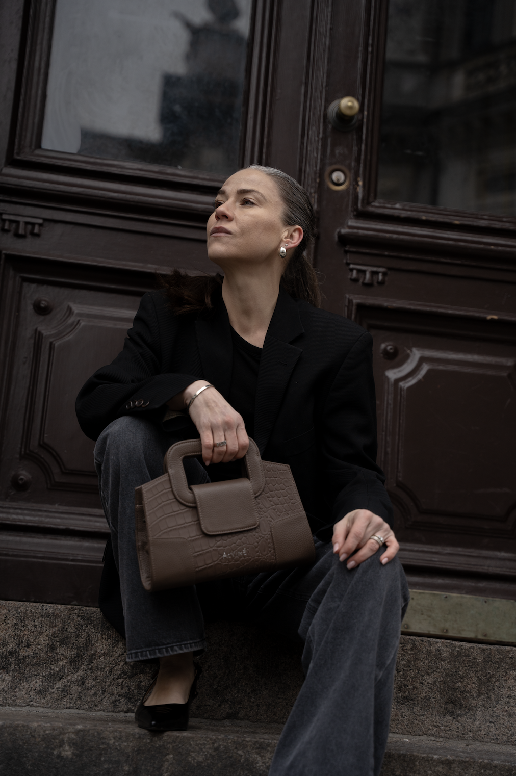 En kvindelig person sidder udenfor foran en trædør, iført en sort blazer og blå jeans, og holder en beige taske.