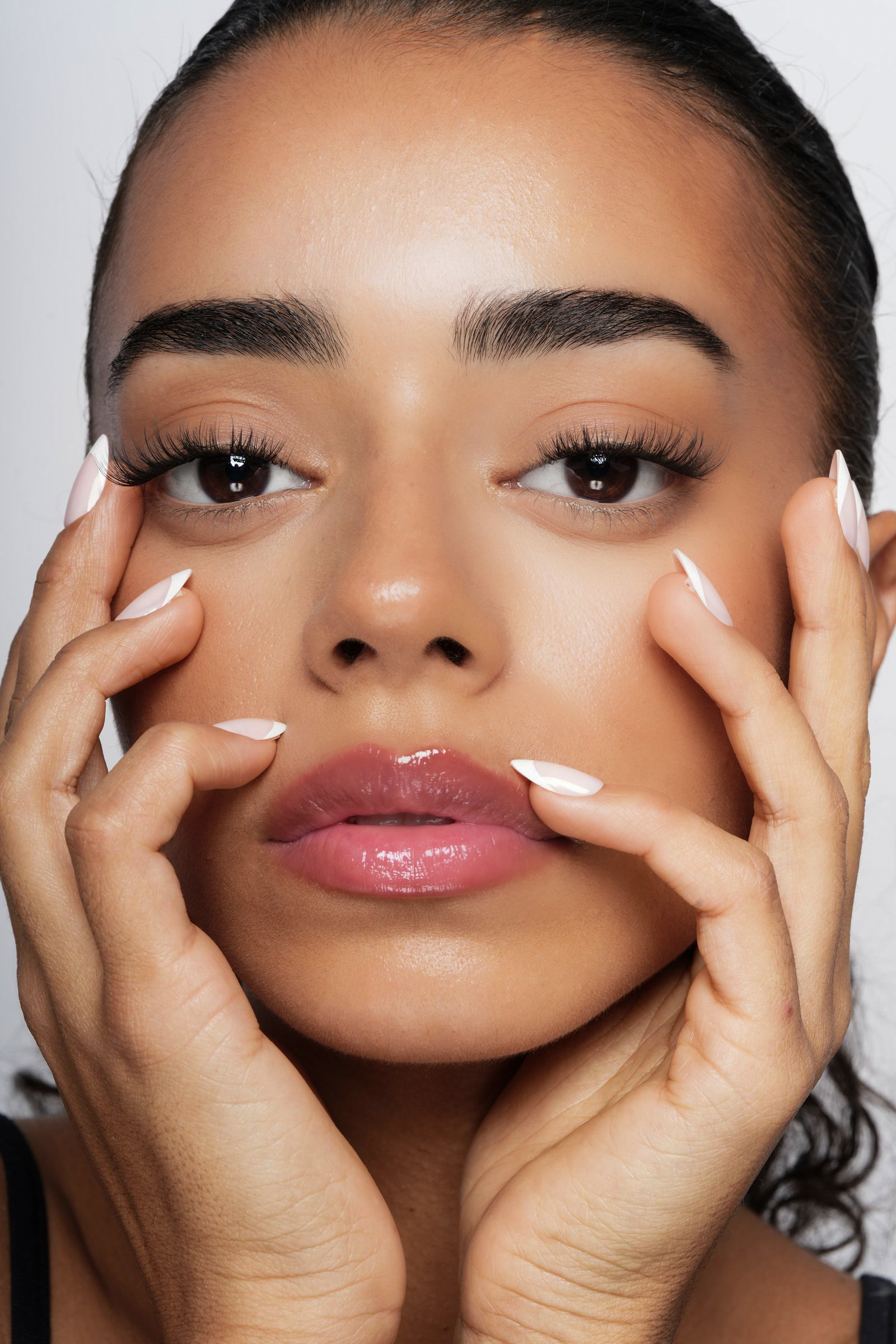 Nærbillede af en kvindes ansigt med fokus på make-up, med hænder ved kinder og hage, lys hud, mørke øjne, velplejede øjenbryn og lyserøde læber.