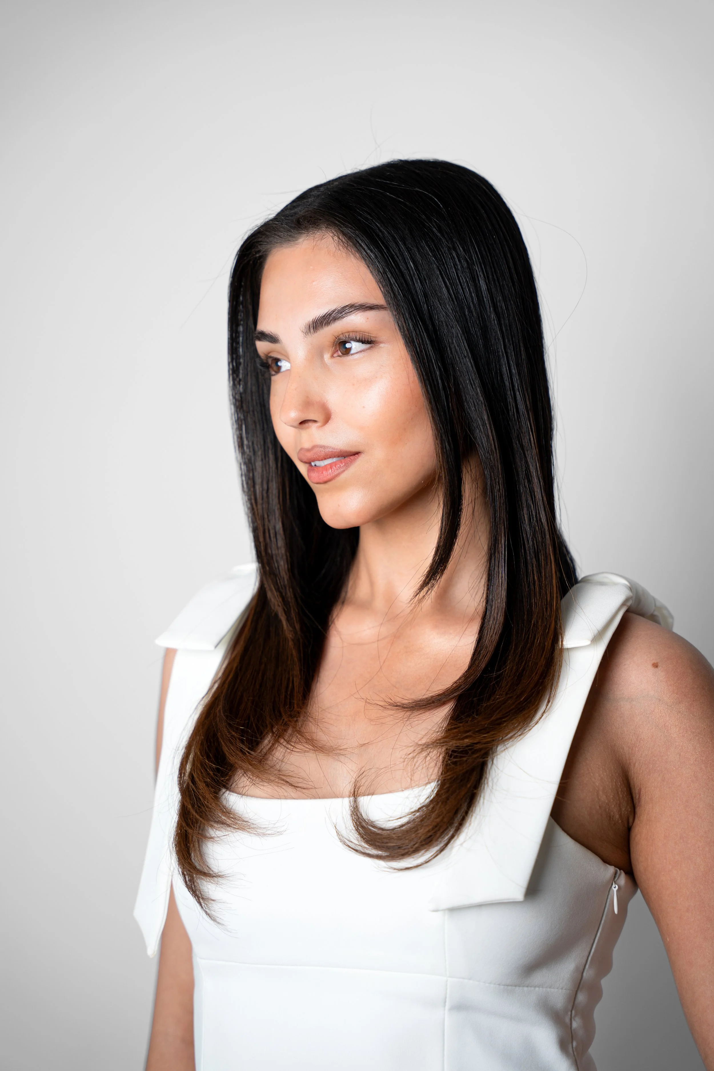 Portræt af en ung kvinde med mørkt hår og lys hud, iført en hvid kjole, set fra siden mod venstre, mod en neutral grå baggrund.