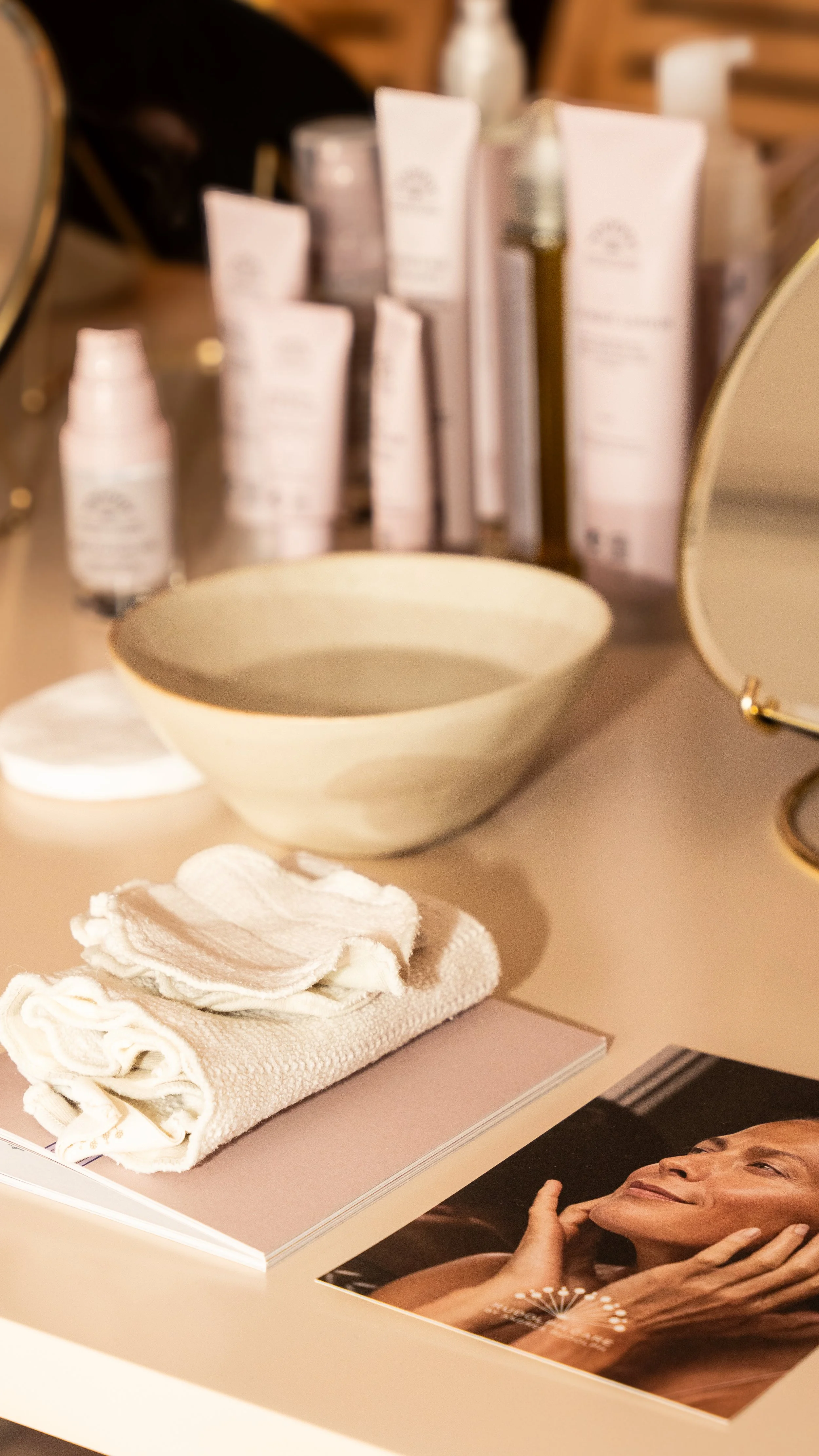 En bordplade med skønhedsprodukter, herunder en stor hvælvet skål, flere rensende produkter i hvide og grå emballager, en skrubbeklude, en vaskedims og et magasin med et billede af en afslappet kvinde.