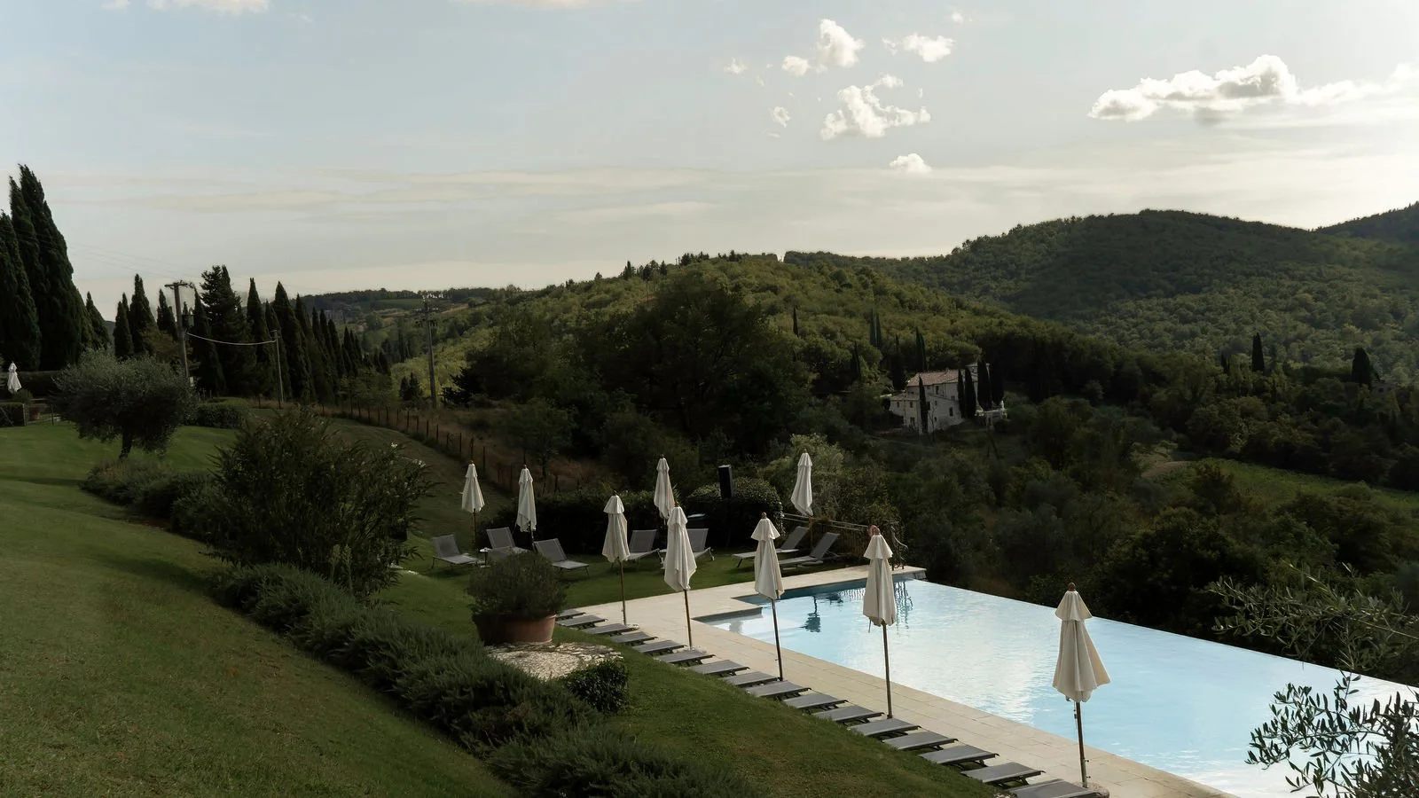 Poolområde med liggestole og parasoller, omgivet af grønne bakker og træer i det italienske landskab.