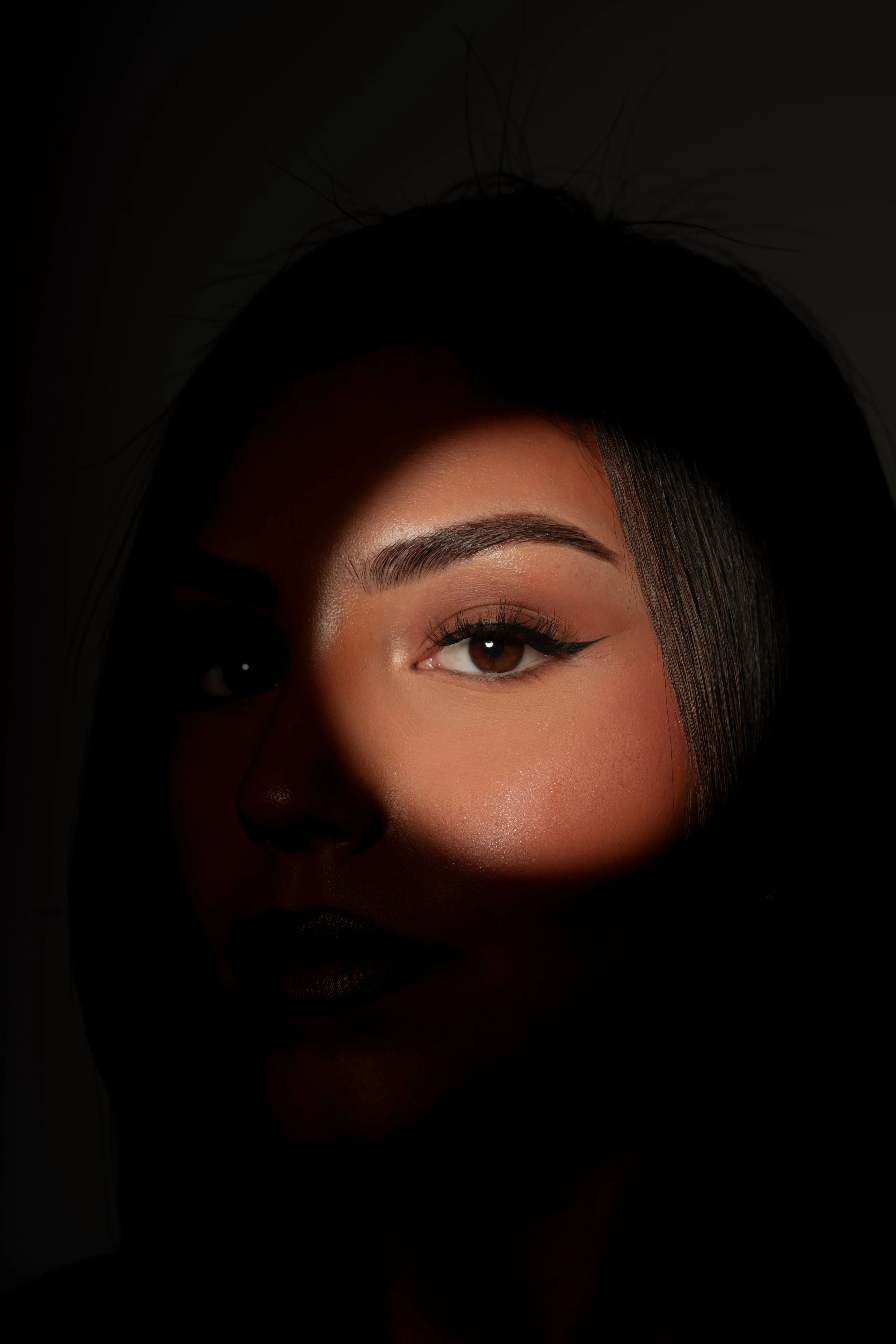 Nærbillede af en kvinde med make-up, hvor kun halvdelen af ansigtet er synligt, oplyst af en lysstribe, med fokus på øje, øjenbryn og læber.