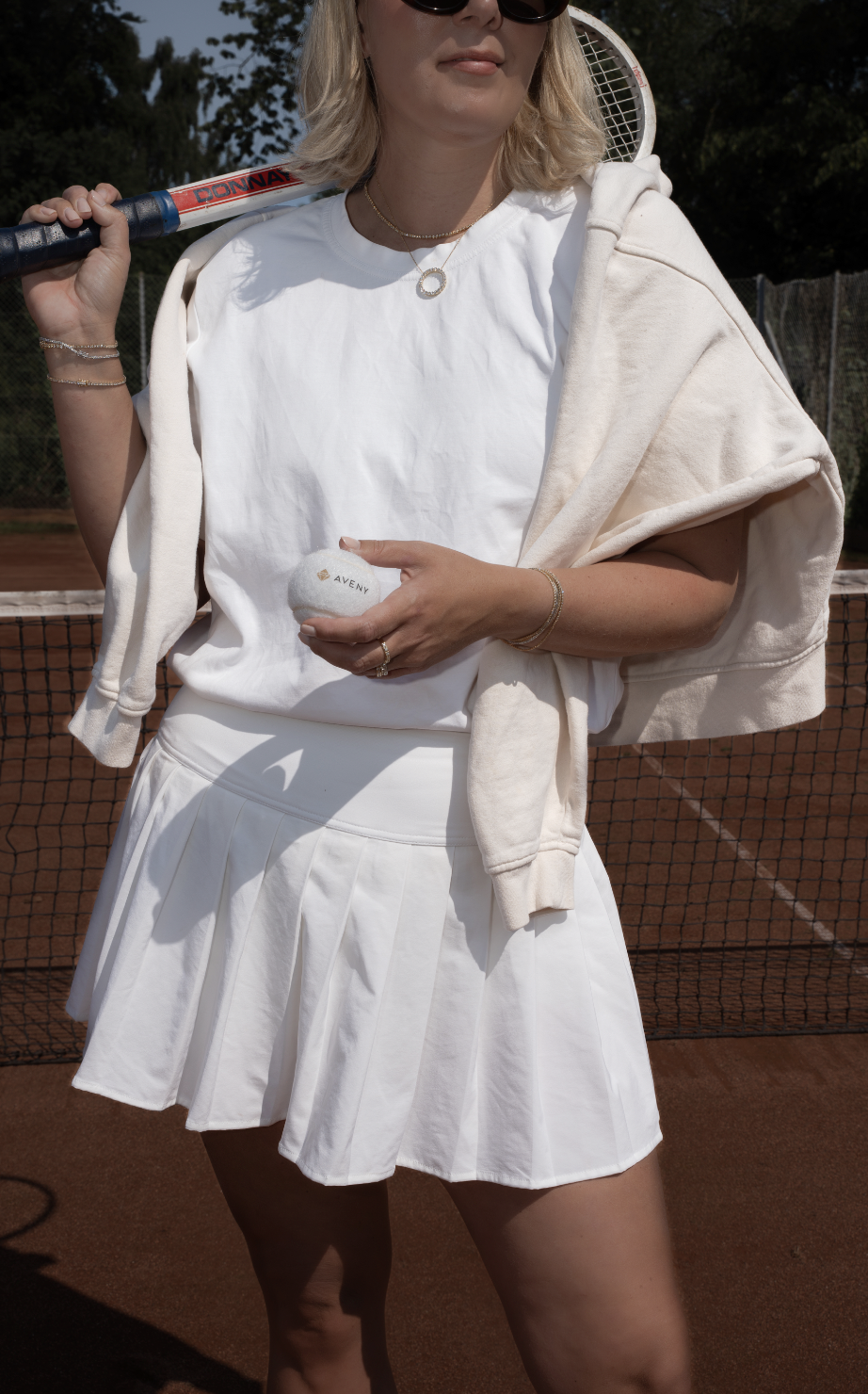 Kvinde på tennisbane iført hvid kjole og solbriller, der holder en tennisbold.