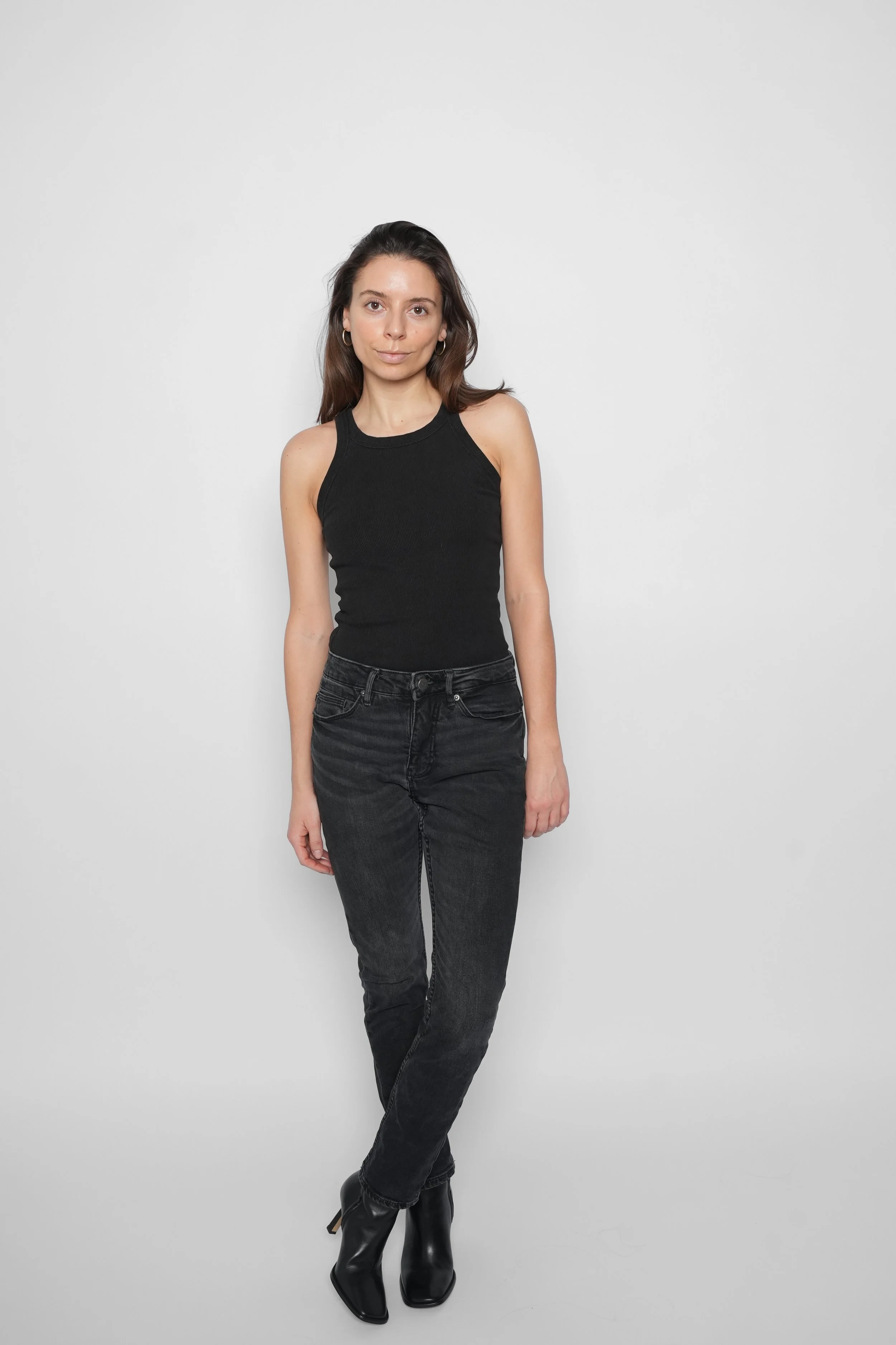 Kvinde i sort tanktop, sorte jeans og sort støvler står mod hvid baggrund.