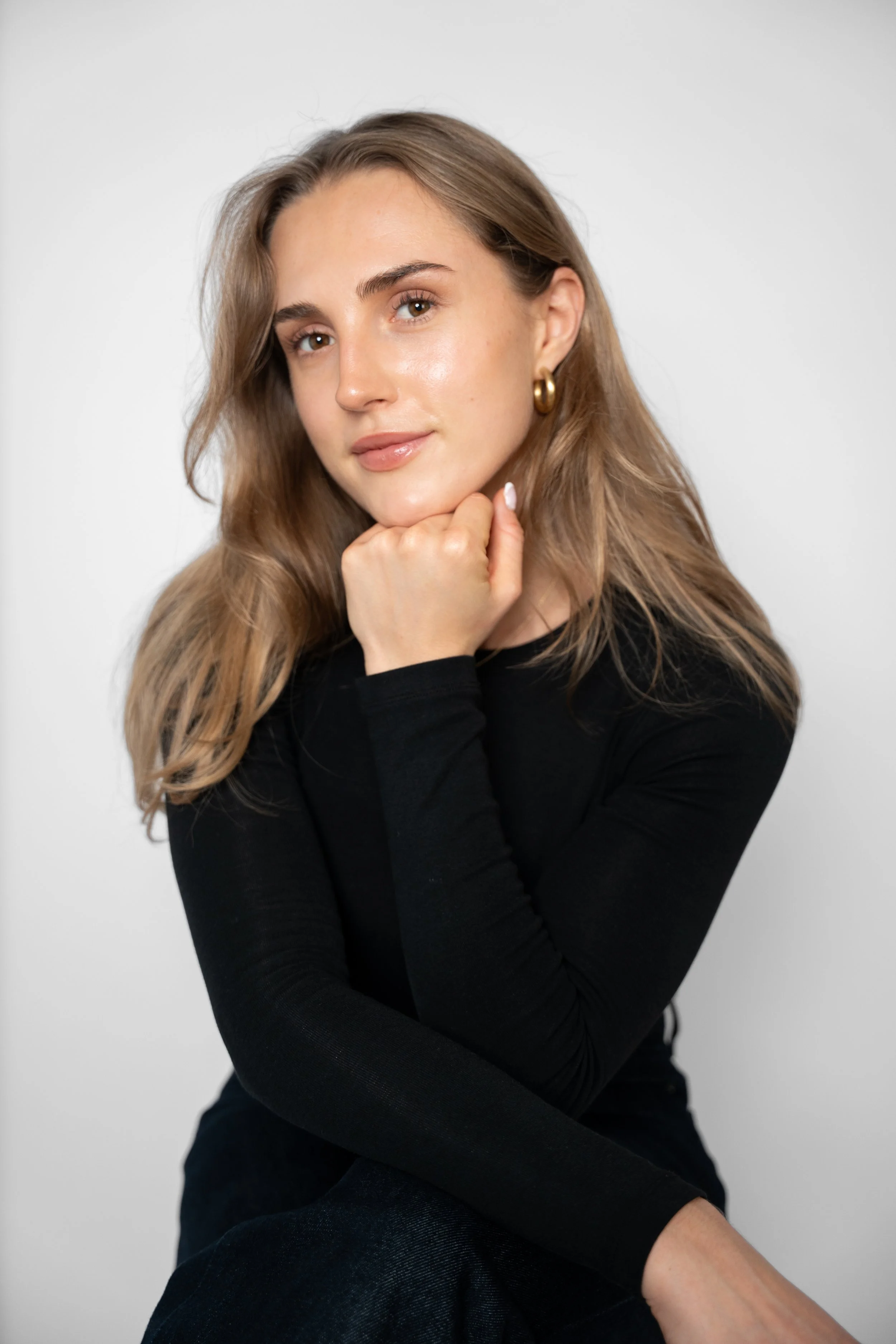 En ung kvinde med lyst brunt hår, iført sort langærmet top og store guldører, sidder med hagen hvilende i hånden foran en grå baggrund.