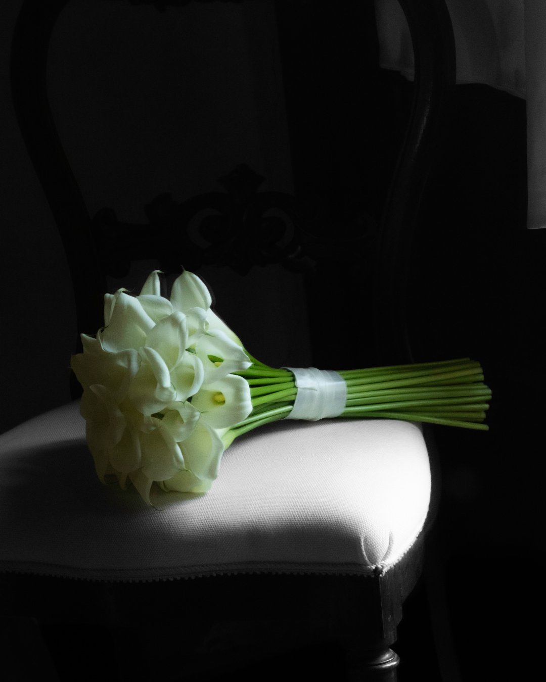 En buket hvide calla-liljer ligger på en hvid stol, med en mørk baggrund