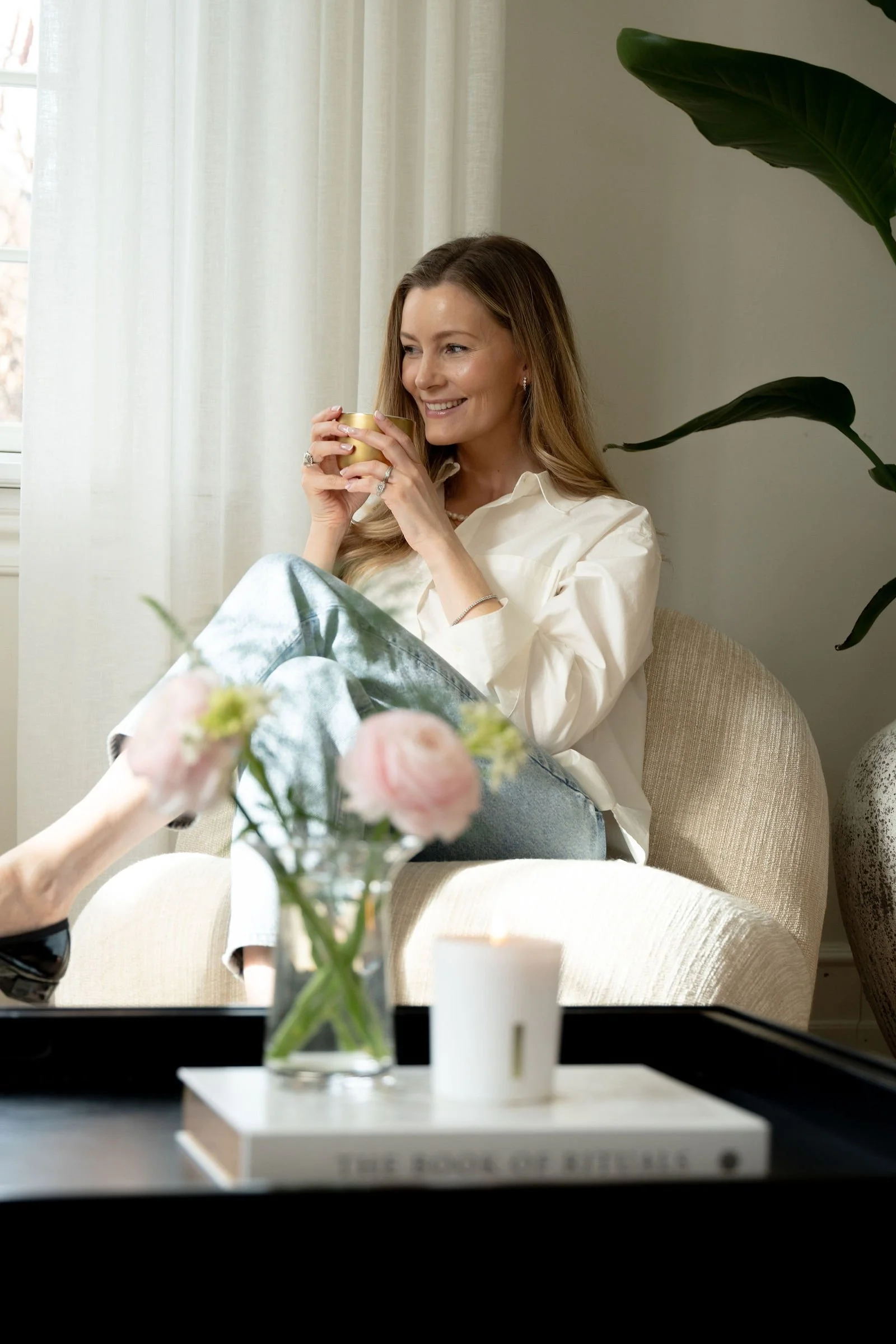 En smilende kvinde sidder i en beige lænestol med benene oppe, holder en kop og nyder et øjeblik. Der er blomster i en vase og en tændt stearinlys på et lavt sort bord foran hende.