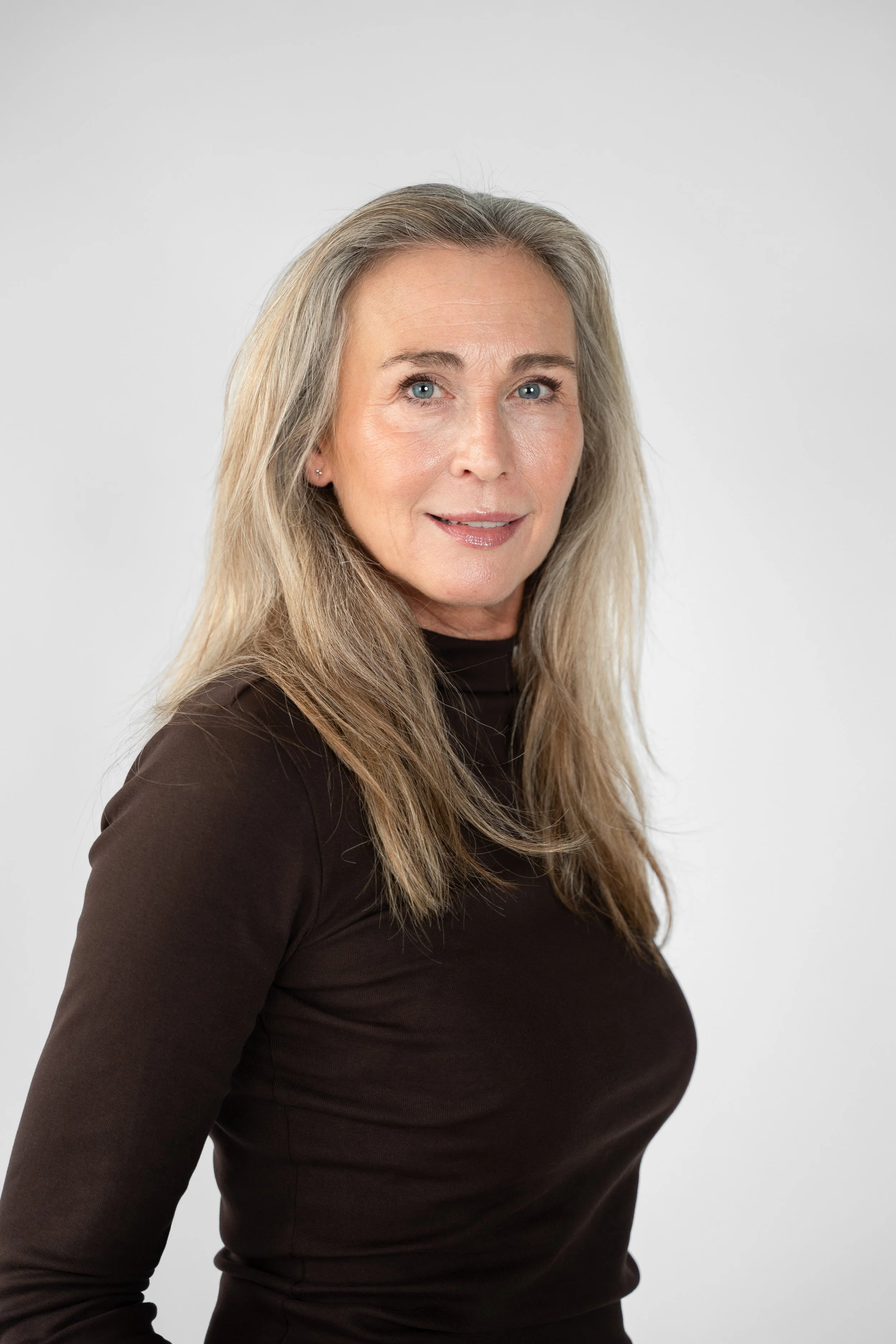 Portrett af en kvinde med blondt, langt hår, iført en sort langærmet top, mod en neutral, lysegrå baggrund.