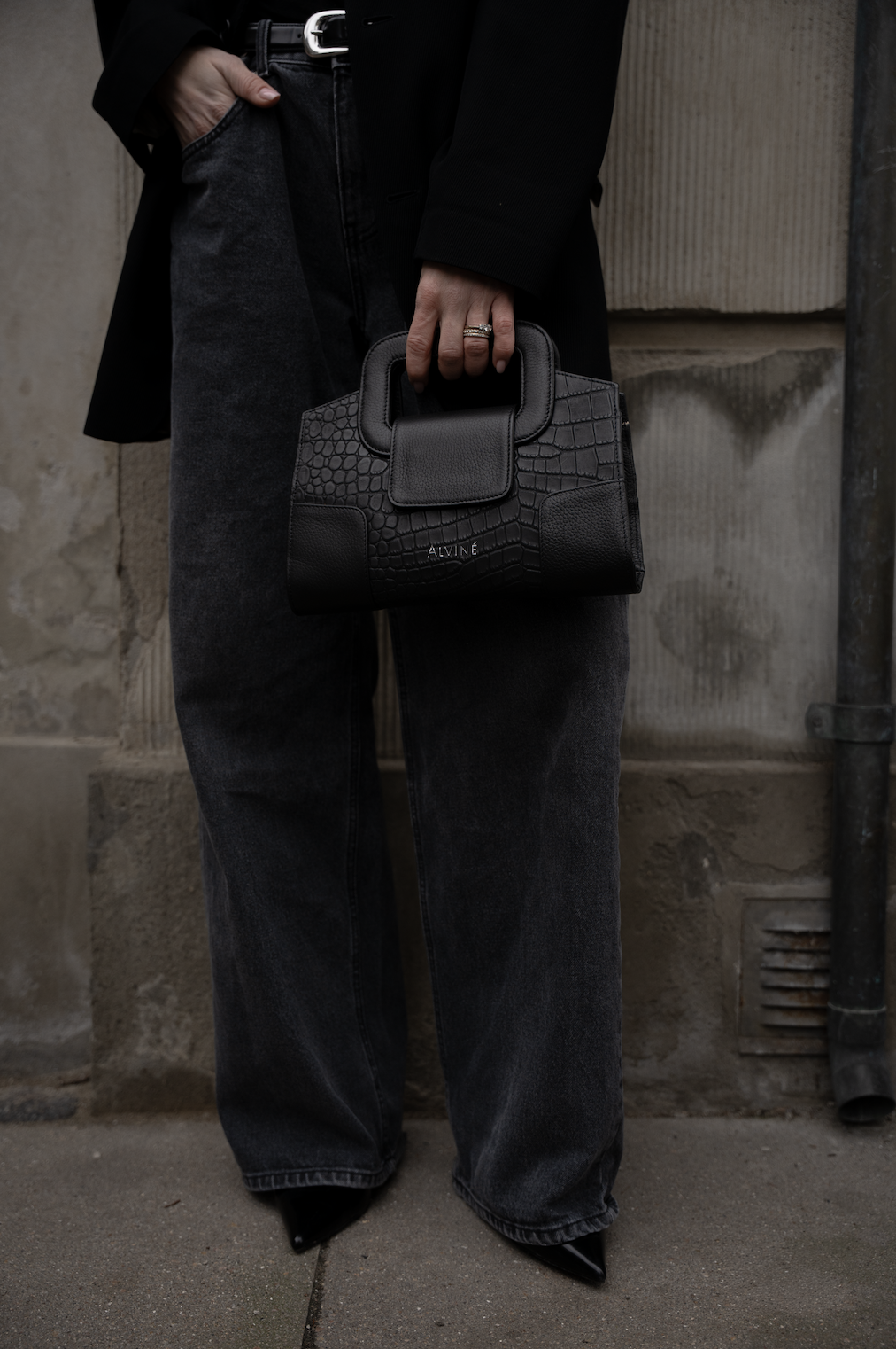 Person der holder en sort taske med mærket ALVINÉ, iført sort jakke, løse sorte jeans og sorte sko. Baggrunden er en murstensvæg.