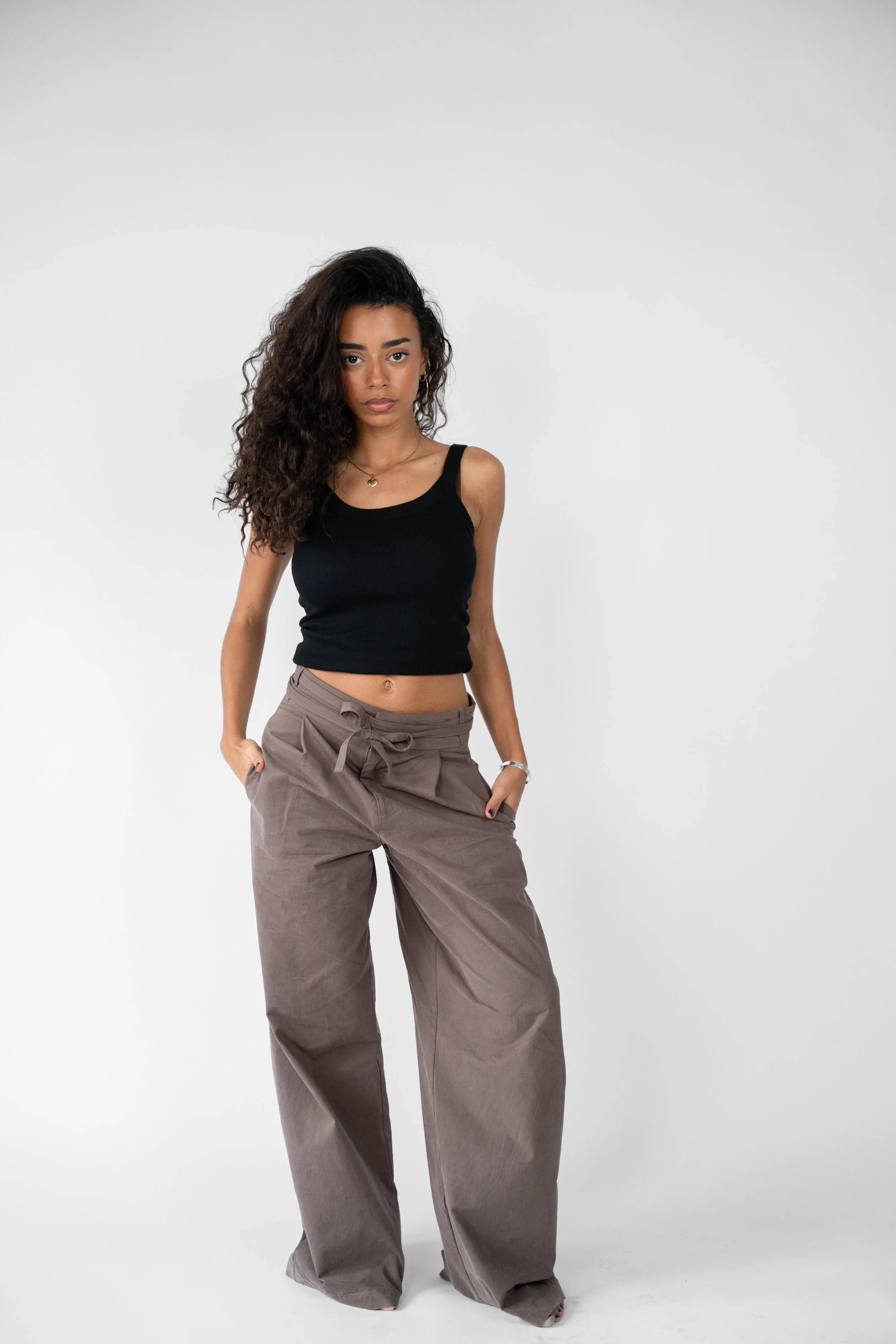 Ung kvinde med curly hår, sort top og bæltebukser, stående med hænder i lommerne mod en hvid baggrund.