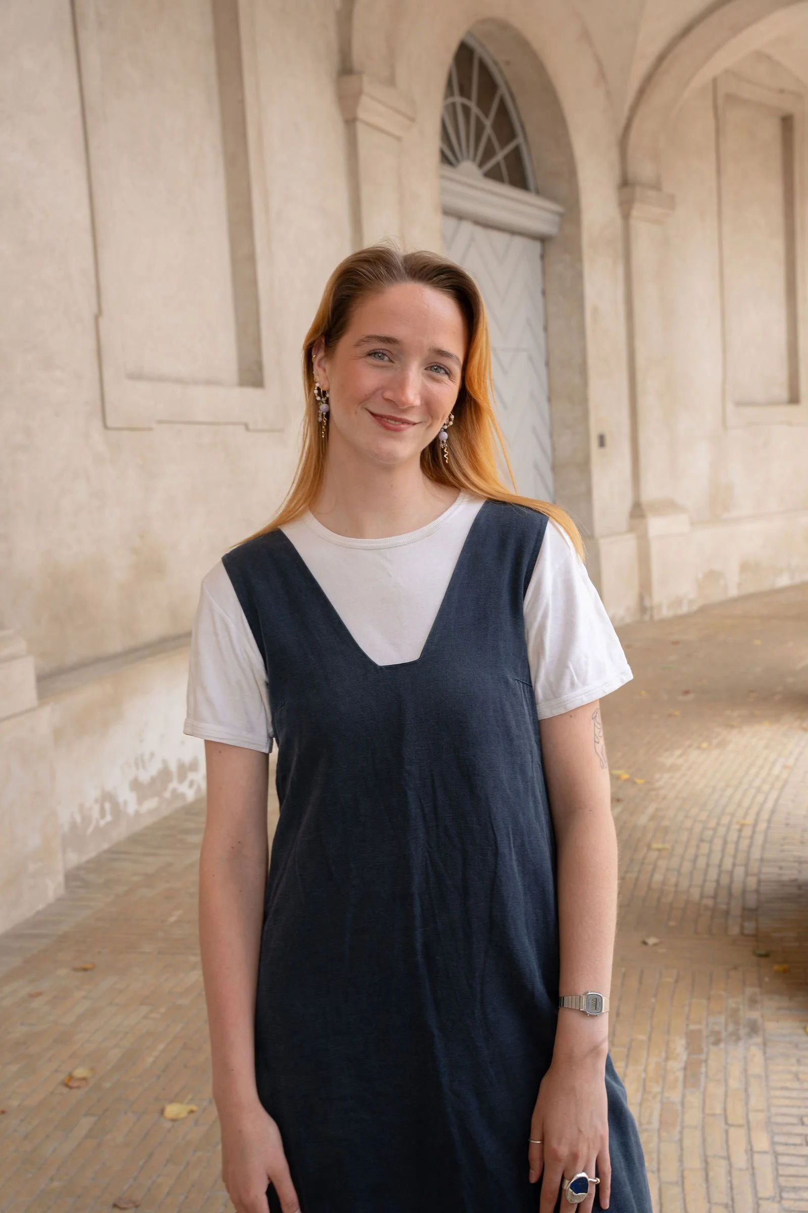 En smilende kvinde med rødt hår, der står udenfor under en bue, iført en hvid T-shirt og en mørkeblå kjole.