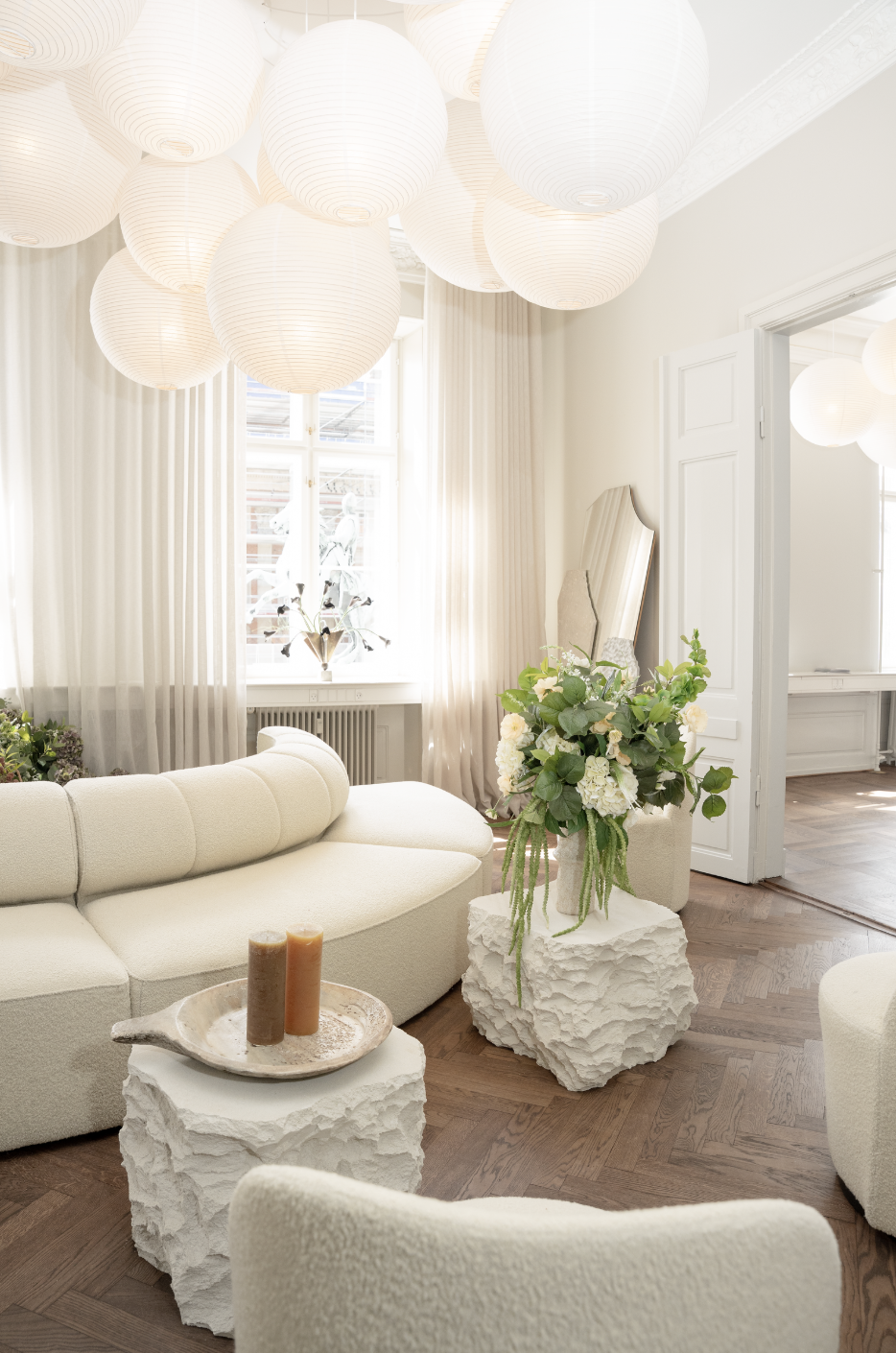 Lyselejet og moderne stue med hvide papirslygter i loftet, hvid sofa, grønne planter med hvide blomster og træbord med stearinlys.