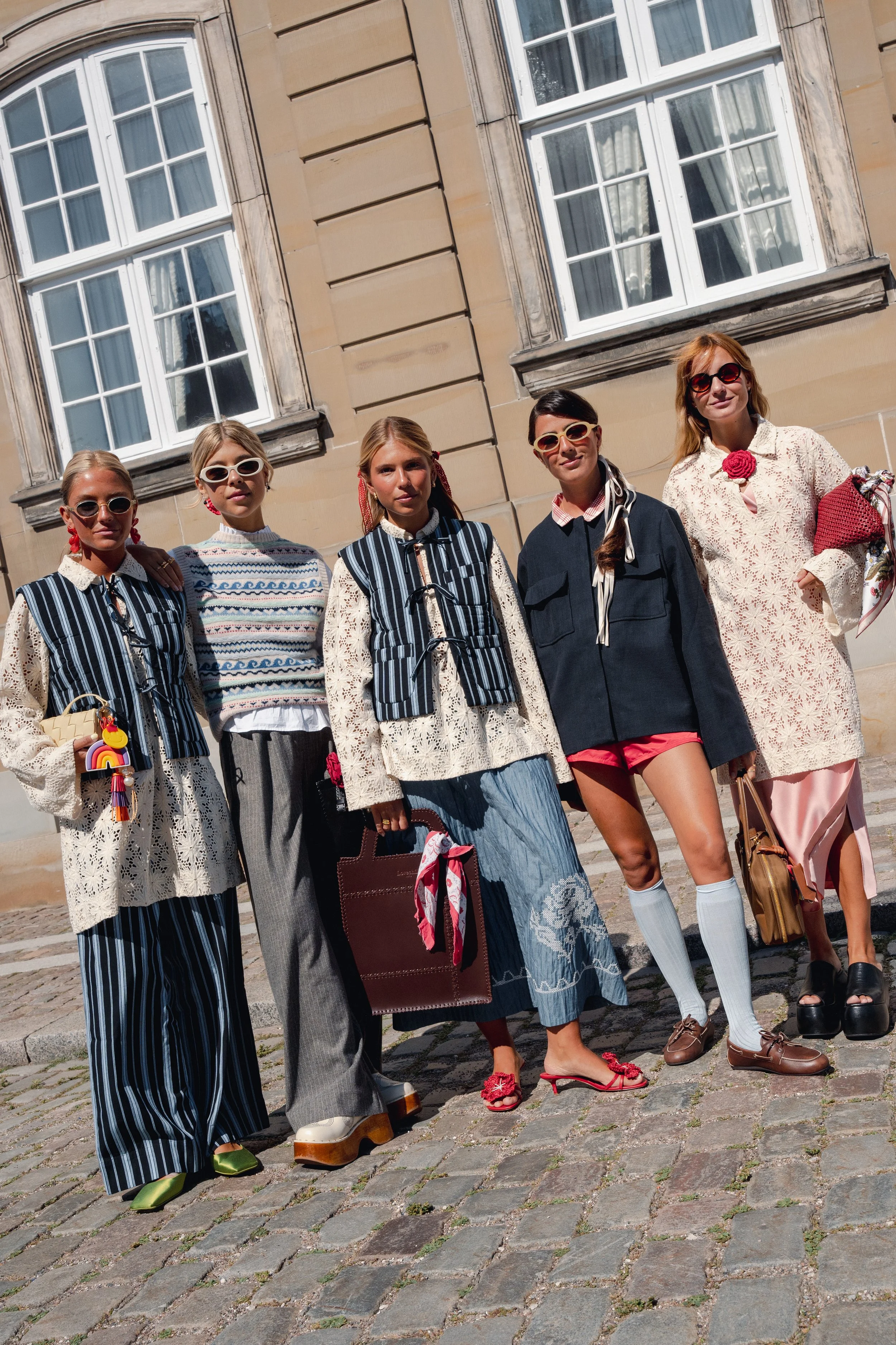 Fem kvinder står foran en bygning med store vinduer, iført farverige og mønstrede tøj, samt solbriller.